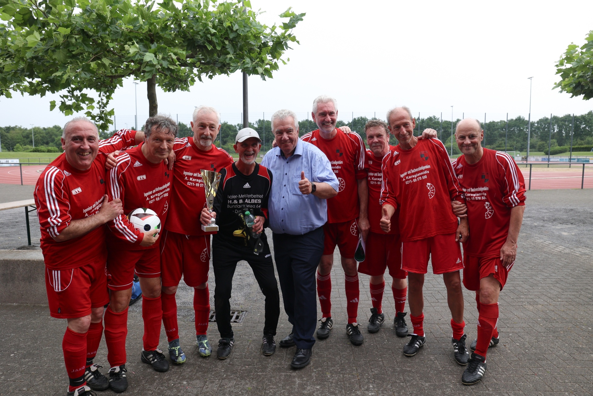 [{"clang_id":1,"value":"Fußballmannschaft in roten Trikots posiert mit Pokalen. Ein Mann in zivil kleidet steht in der Mitte der Gruppe. Im Hintergrund Sportplatz und Bäume."}]