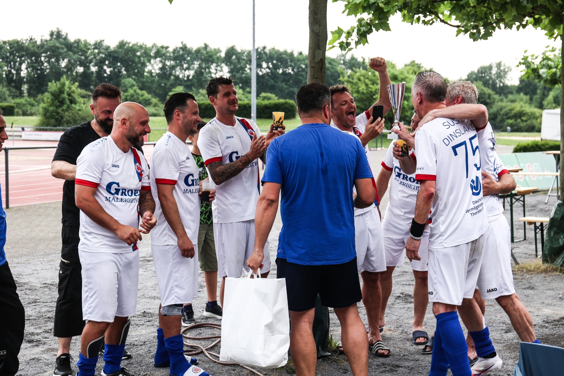 [{"clang_id":1,"value":"Fußballspieler in weißen und blauen Trikots feiern mit einem Mann in blauem Shirt und einer Trophäe am Spielfeldrand. Im Hintergrund Bäume und Zuschauer."}]