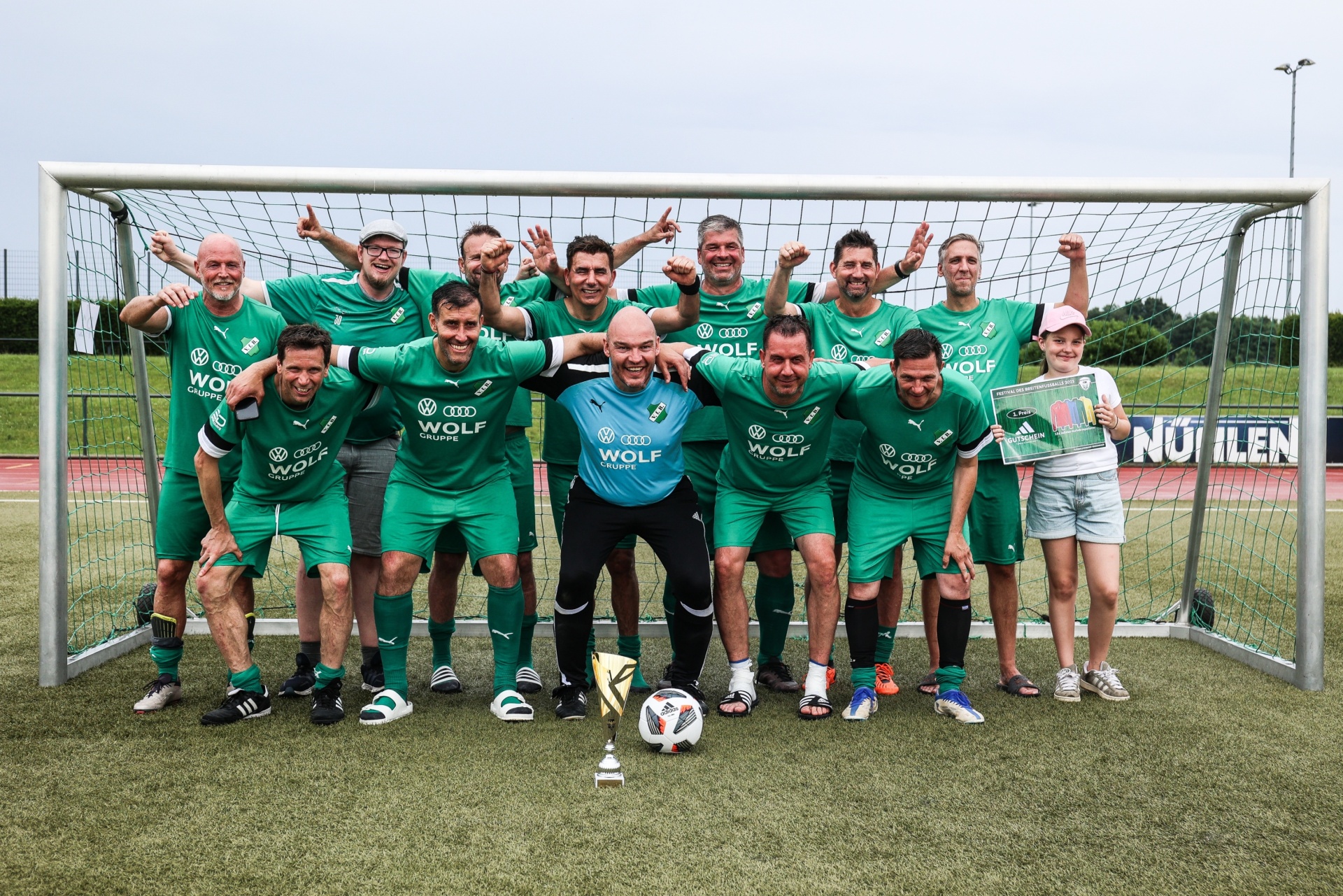 [{"clang_id":1,"value":"Fußballmannschaft in grünen Trikots posiert jubelnd vor einem Fußballtor auf einem Rasenplatz. Sie halten eine Trophäe und eine Siegerfahne."}]