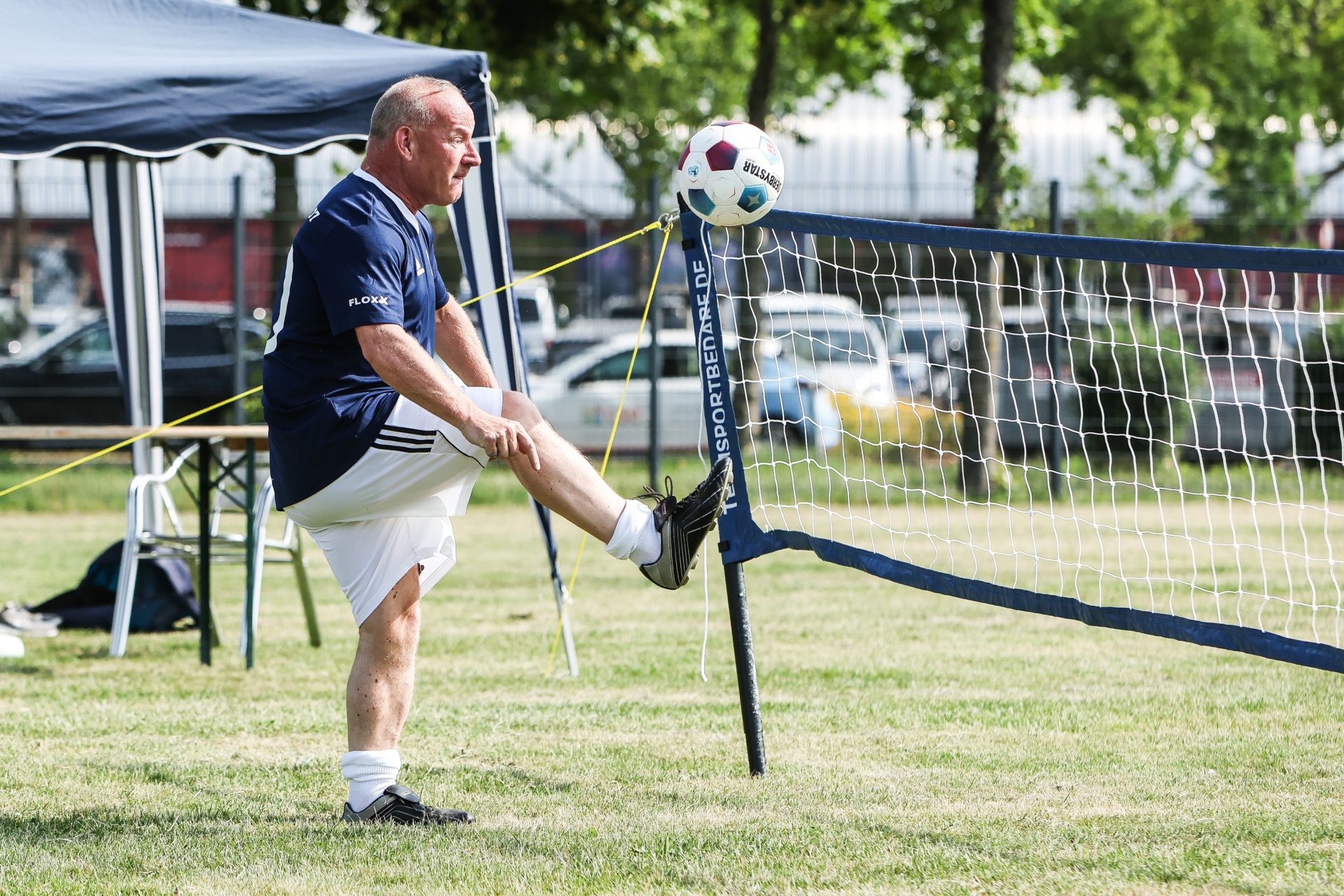 [{"clang_id":1,"value":"Mann kickt Ball am Volleyballnetz auf grünem Rasen. Im Hintergrund ein Zelt und Bäume."}]