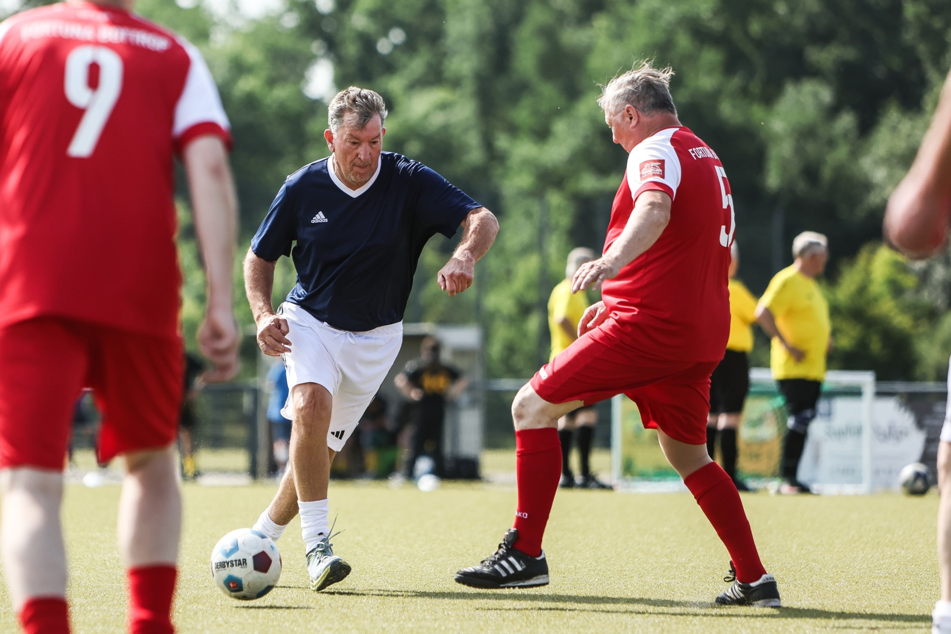 [{"clang_id":1,"value":"Zwei ältere Männer spielen Fußball auf einem grünen Rasenplatz. Links ein Spieler in Dunkelblau, rechts in Rot. Im Hintergrund weitere Spieler und Zuschauer."}]