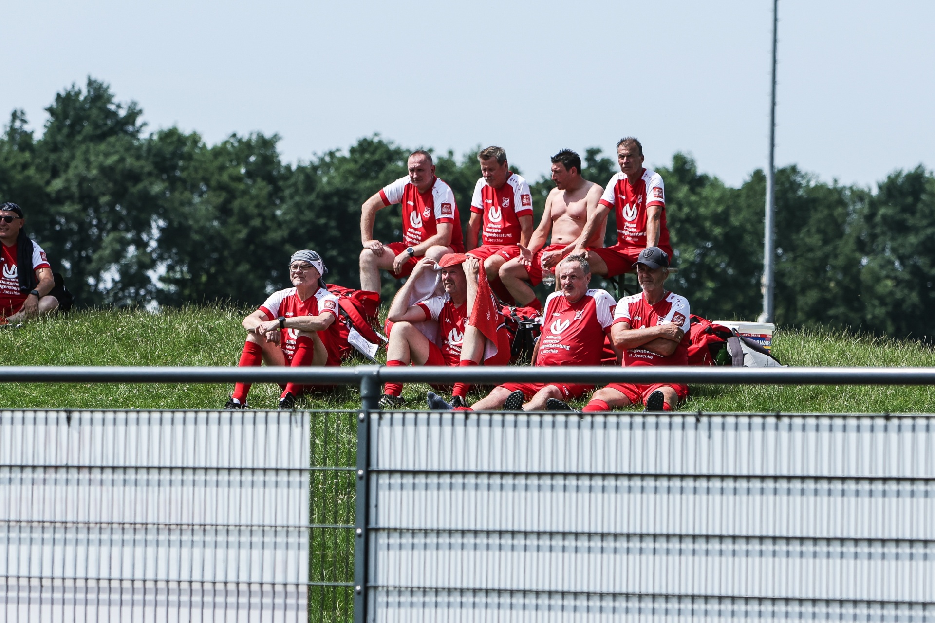 [{"clang_id":1,"value":"Rugby-Mannschaft sitzt und lehnt an einer Metallbarriere am Spielfeldrand. Spieler tragen rote und weiße Trikots, einige sind halbnackt. Im Hintergrund Bäume und Himmel."}]