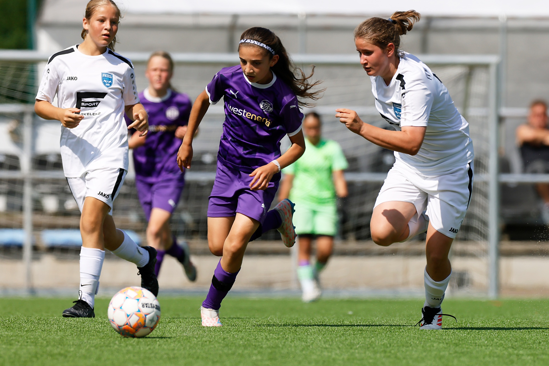 [{"clang_id":1,"value":"Fußballspiel: Zwei junge Spielerinnen in weißen und violetten Trikots dribbeln den Ball auf grünem Rasen, weitere Spieler im Hintergrund."}]