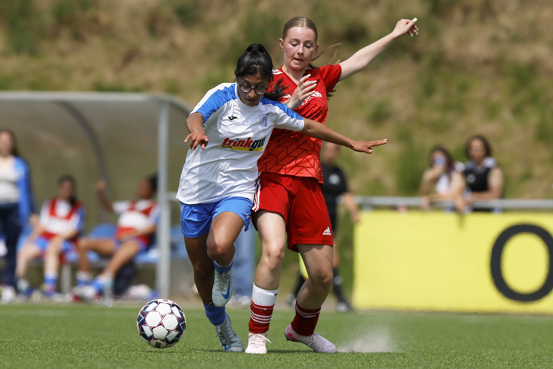 [{"clang_id":1,"value":"Zwei Fußballspielerinnen kämpfen um den Ball auf einem grünen Rasenplatz. Links trägt eine Spielerin blaues Trikot, rechts rotes. Im Hintergrund Zuschauer und ein Punktezahlen-Schild mit der \"0\"."}]
