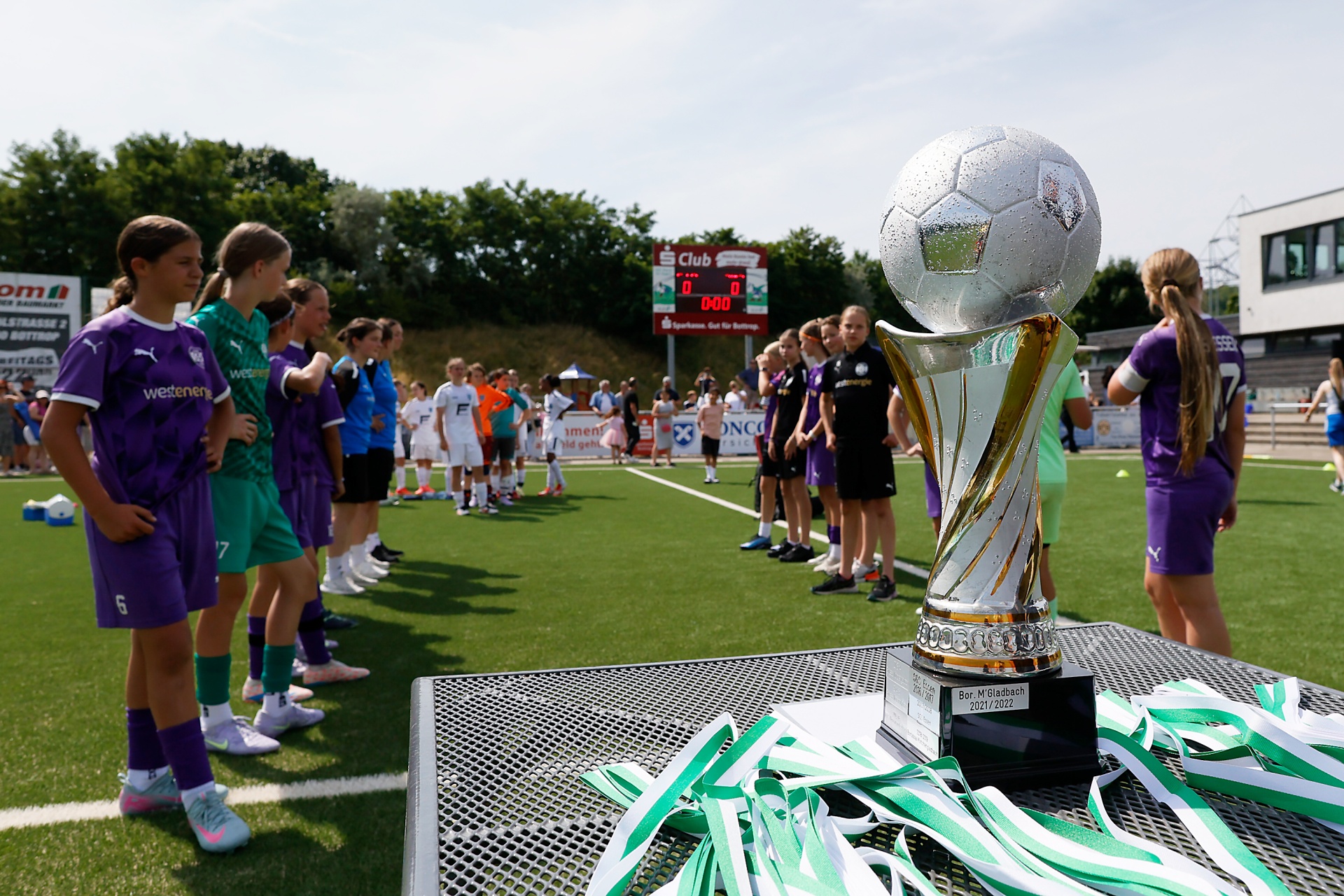[{"clang_id":1,"value":"Fußballturnier: Jugendmannschaft in violetten Trikots steht vor einer silbernen Pokal mit Fußball-Dekoration und grün-weißen Bändern auf dem Rasen. Im Hintergrund Zuschauer und Anzeigetafel."}]