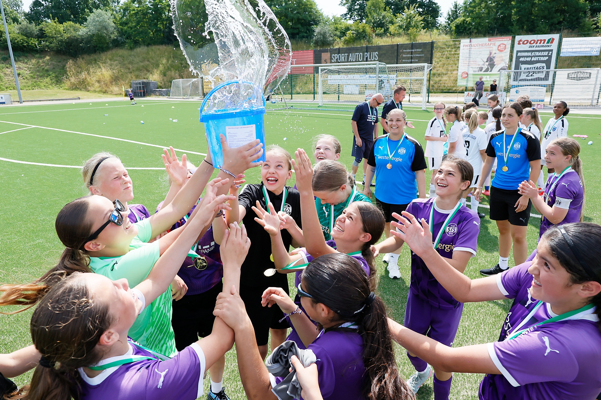 [{"clang_id":1,"value":"Fußballmannschaft jubelt auf grünem Rasen. Spielerinnen in violetten Trikots werfen sich mit Wasser aus einem Eimer zu, Medaillen um den Hals. Hintergrund: Zuschauer und Werbebanner."}]