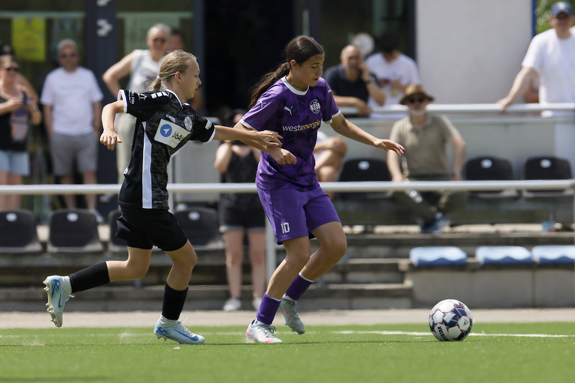 [{"clang_id":1,"value":"Zwei Fußballspielerinnen im Spiel. Links schwarz-weiß gestreiftes Trikot, rechts lilafarbenes Trikot mit Nummer 10. Rasenplatz, Zuschauer im Hintergrund."}]