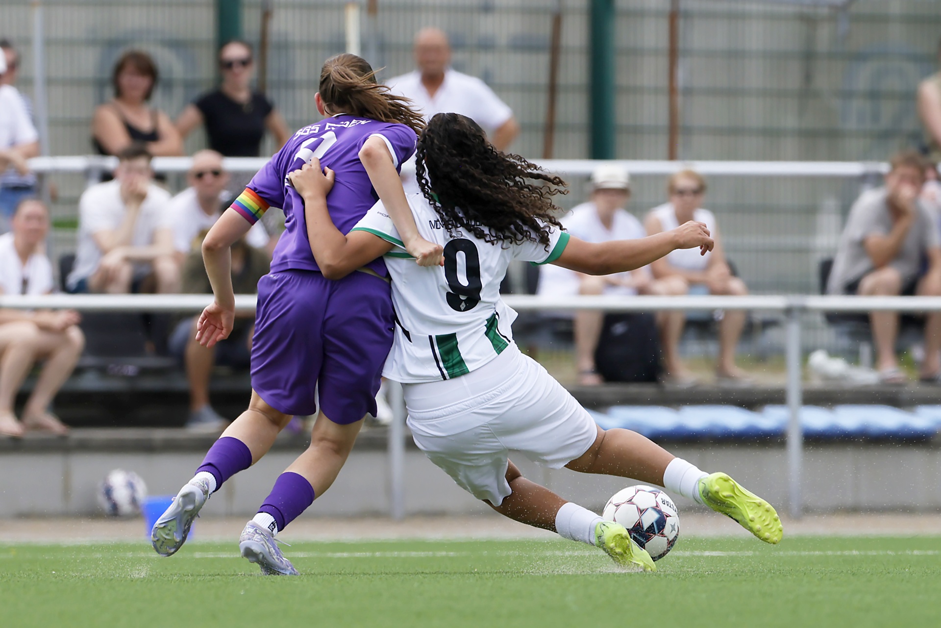 [{"clang_id":1,"value":"Zwei Fußballspielerinnen im Spiel. Eine trägt violettes Trikot, die andere grün-weißes mit Nummer 9. Im Hintergrund Zuschauer auf einer Tribüne."}]