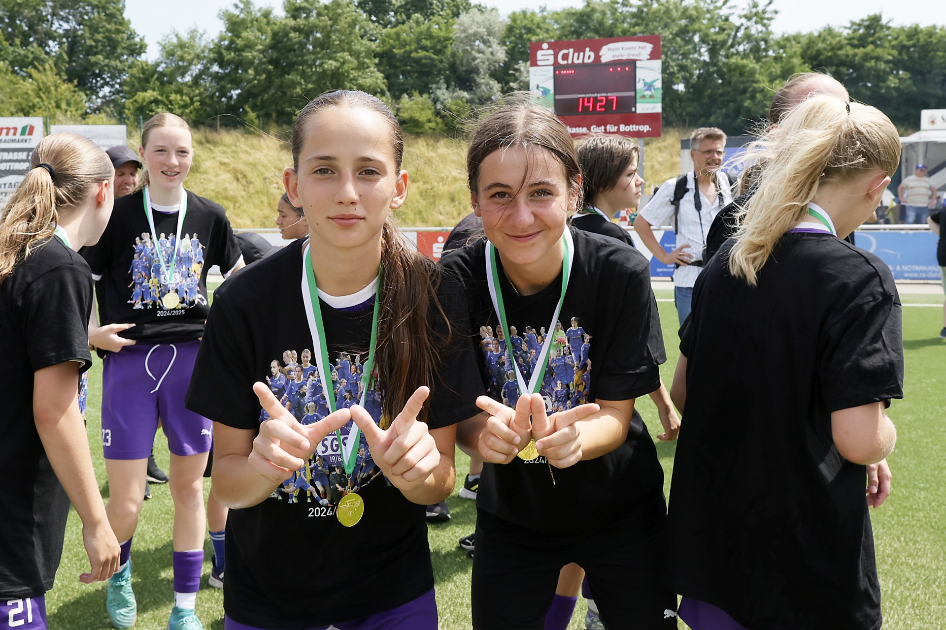[{"clang_id":1,"value":"Zwei Mädchen mit Medaillen um den Hals zeigen Daumen hoch. Im Hintergrund weitere Sportlerinnen auf einem Fußballfeld."}]