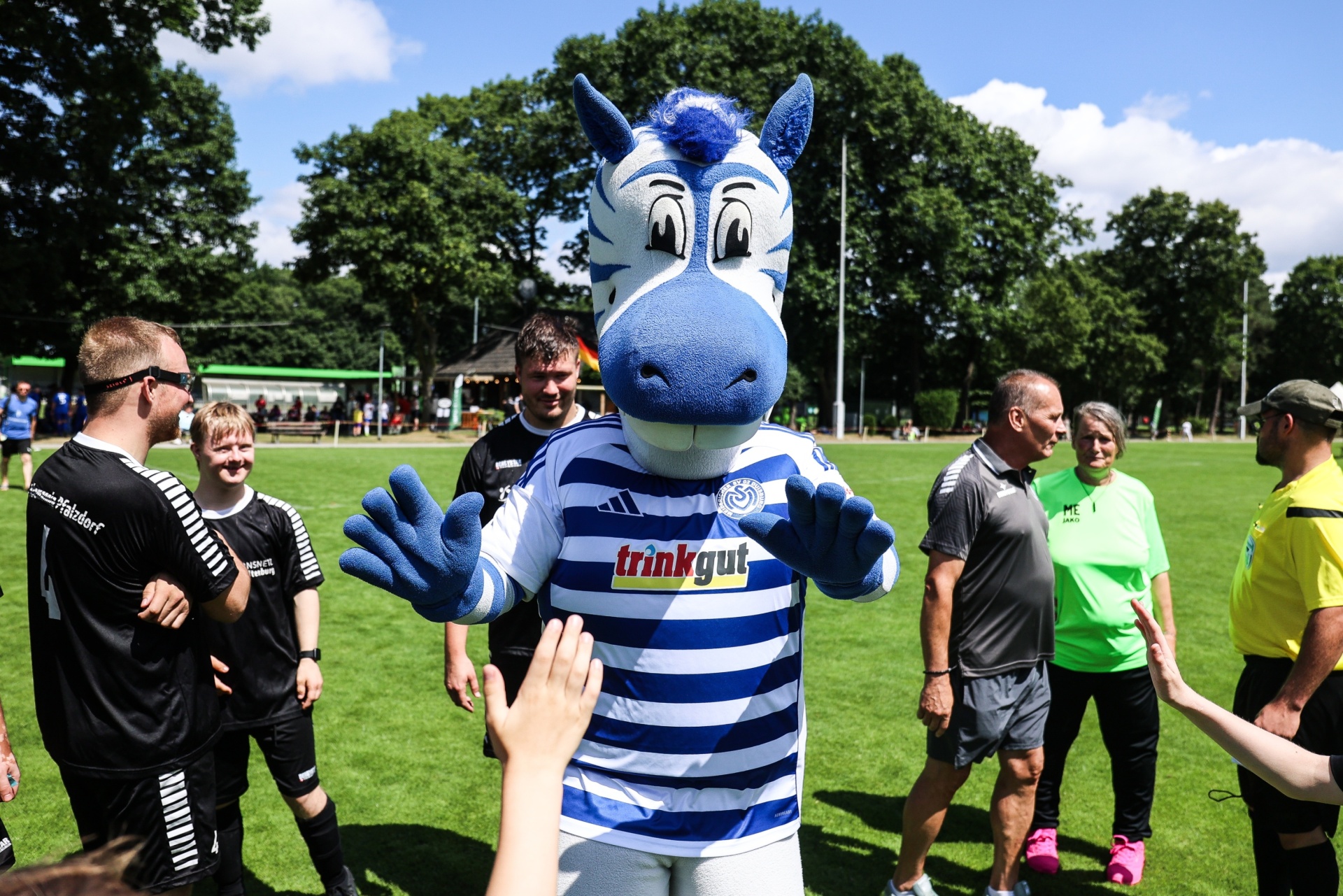 [{"clang_id":1,"value":"Fußballplatz: Blaue Drachen-Mascotte umarmt Spieler in schwarz-weißen Trikots. Im Hintergrund Zuschauer und Bäume bei sonnigem Wetter."}]