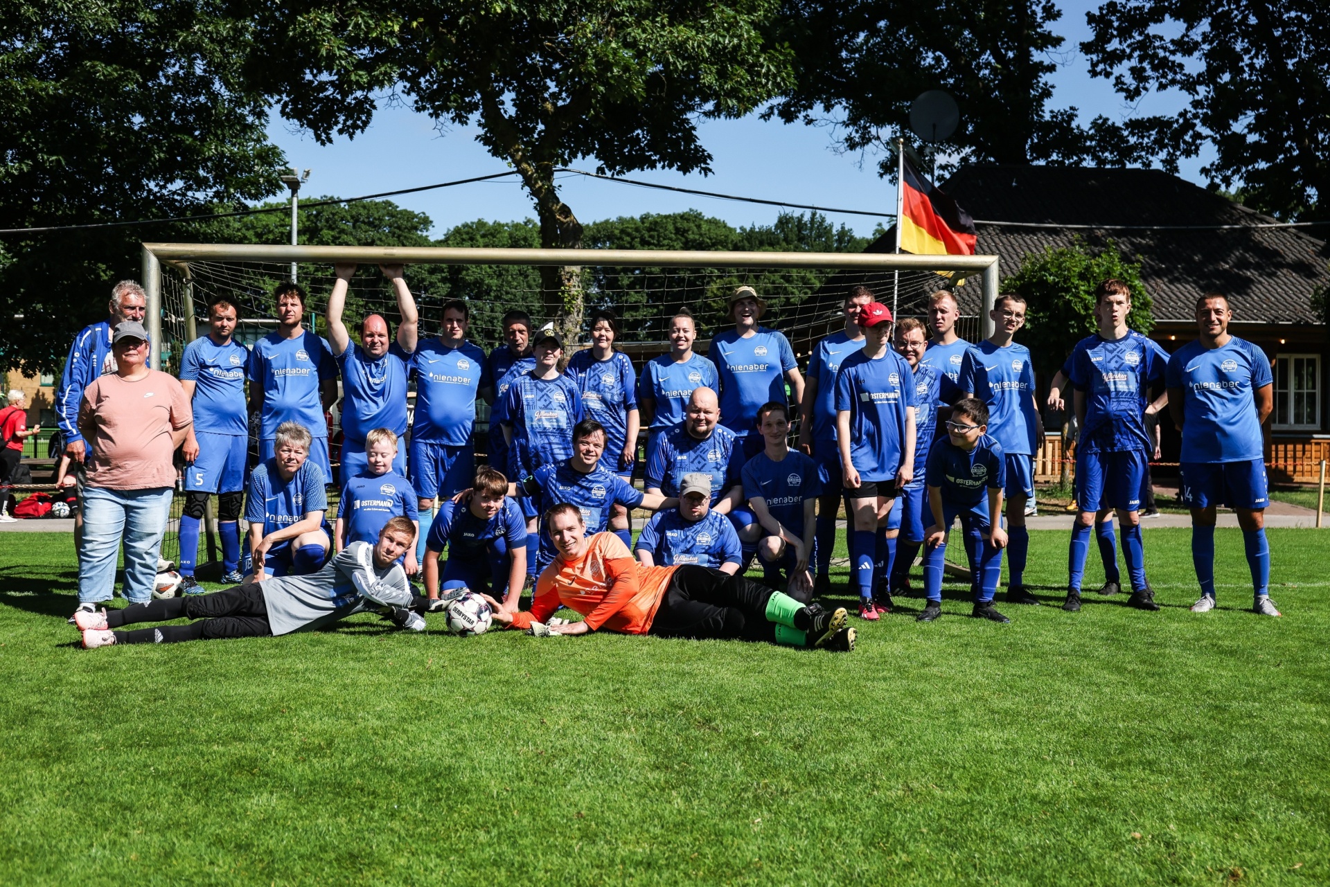 [{"clang_id":1,"value":"Fußballmannschaft in blauen Trikots posiert auf Grün. Im Hintergrund Deutschlandflagge und Fußballtor. Einzelne Personen sitzen oder knien."}]