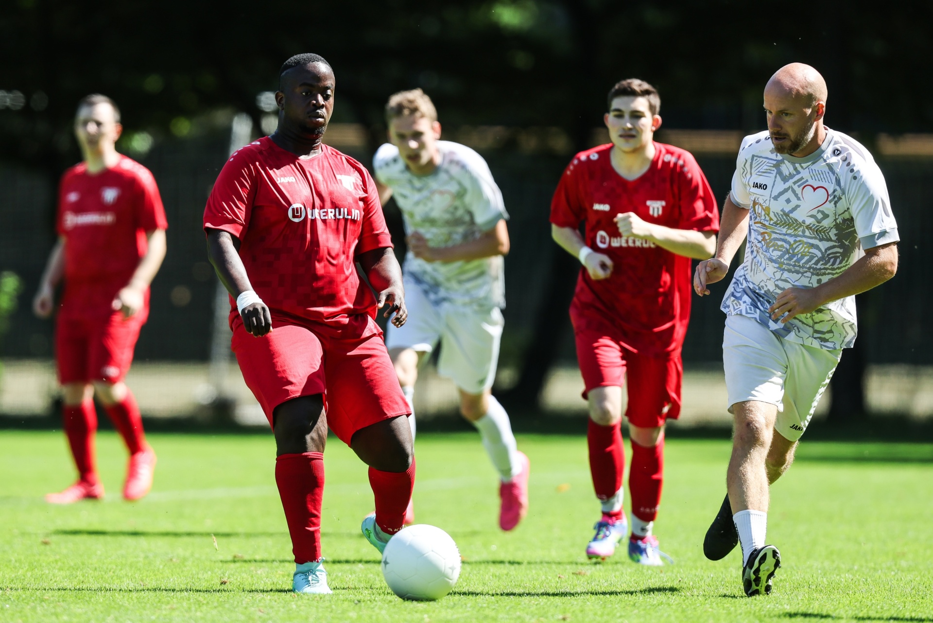 [{"clang_id":1,"value":"Fußballspiel: Spieler in roten und weißen Trikots auf grünem Rasen. Ein Spieler dribbelt den Ball, andere laufen im Hintergrund."}]