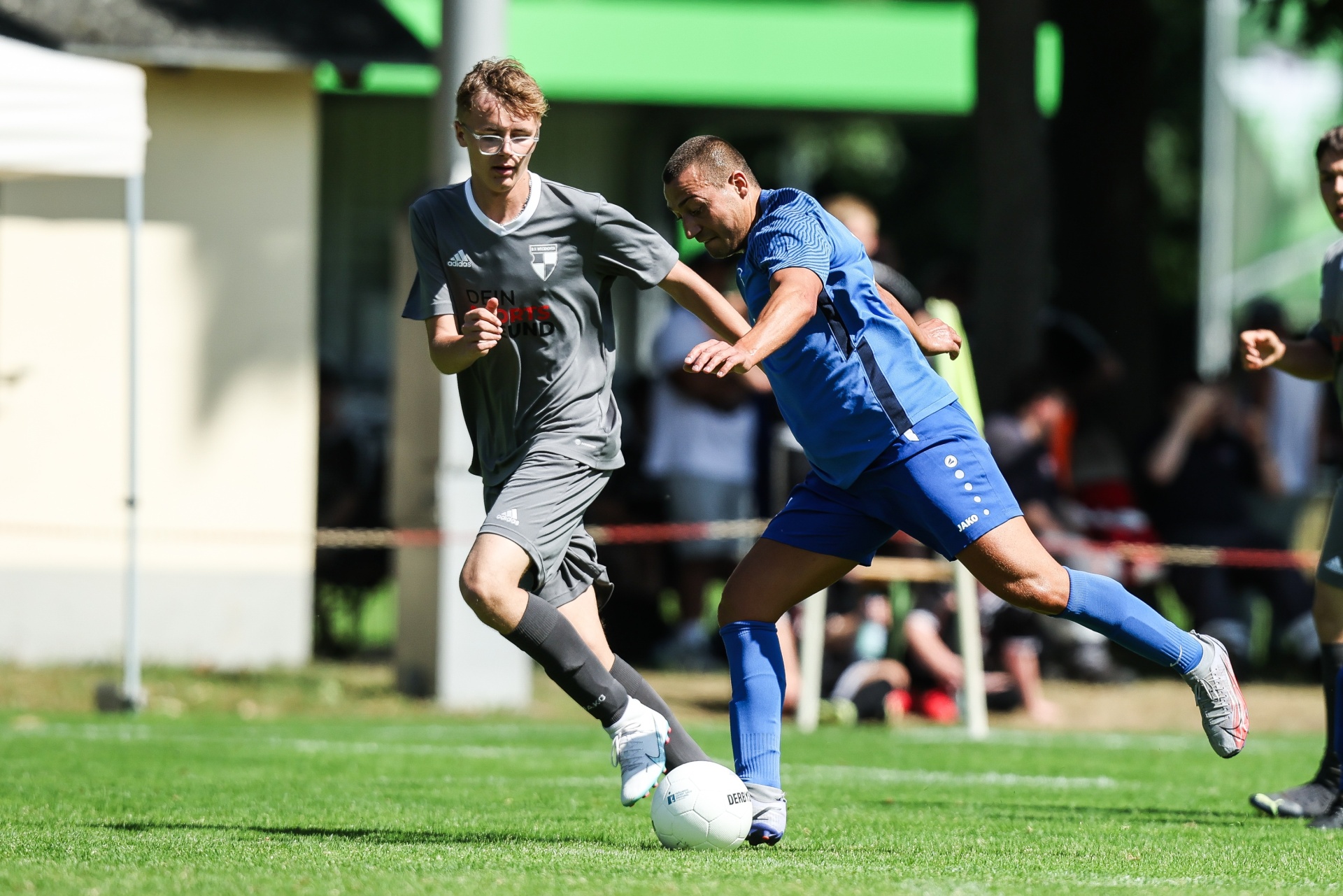 [{"clang_id":1,"value":"Zwei Fußballspieler im Wettkampf. Links ein Spieler mit grauem Trikot, rechts ein Spieler mit blauem Trikot, beide am Ball. Hinter ihnen ist ein Fußballfeld und eine Zuschauertribüne zu sehen."}]