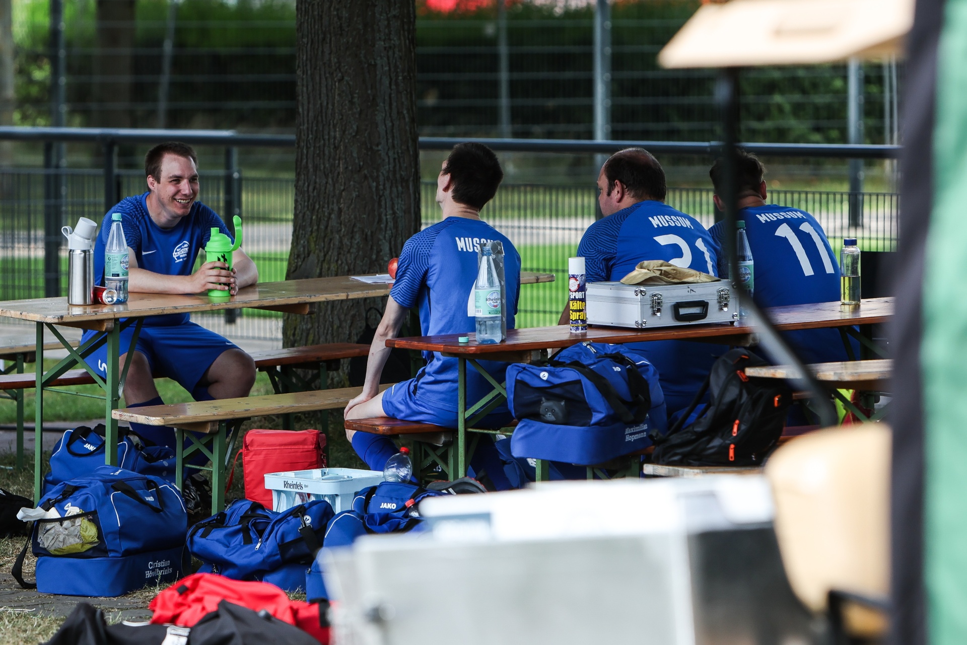[{"clang_id":1,"value":"Fußballspieler sitzen an Picknicktischen im Freien. Sie tragen blaue Trikots und Shorts. Sporttaschen und Wasserflaschen stehen dabei."}]