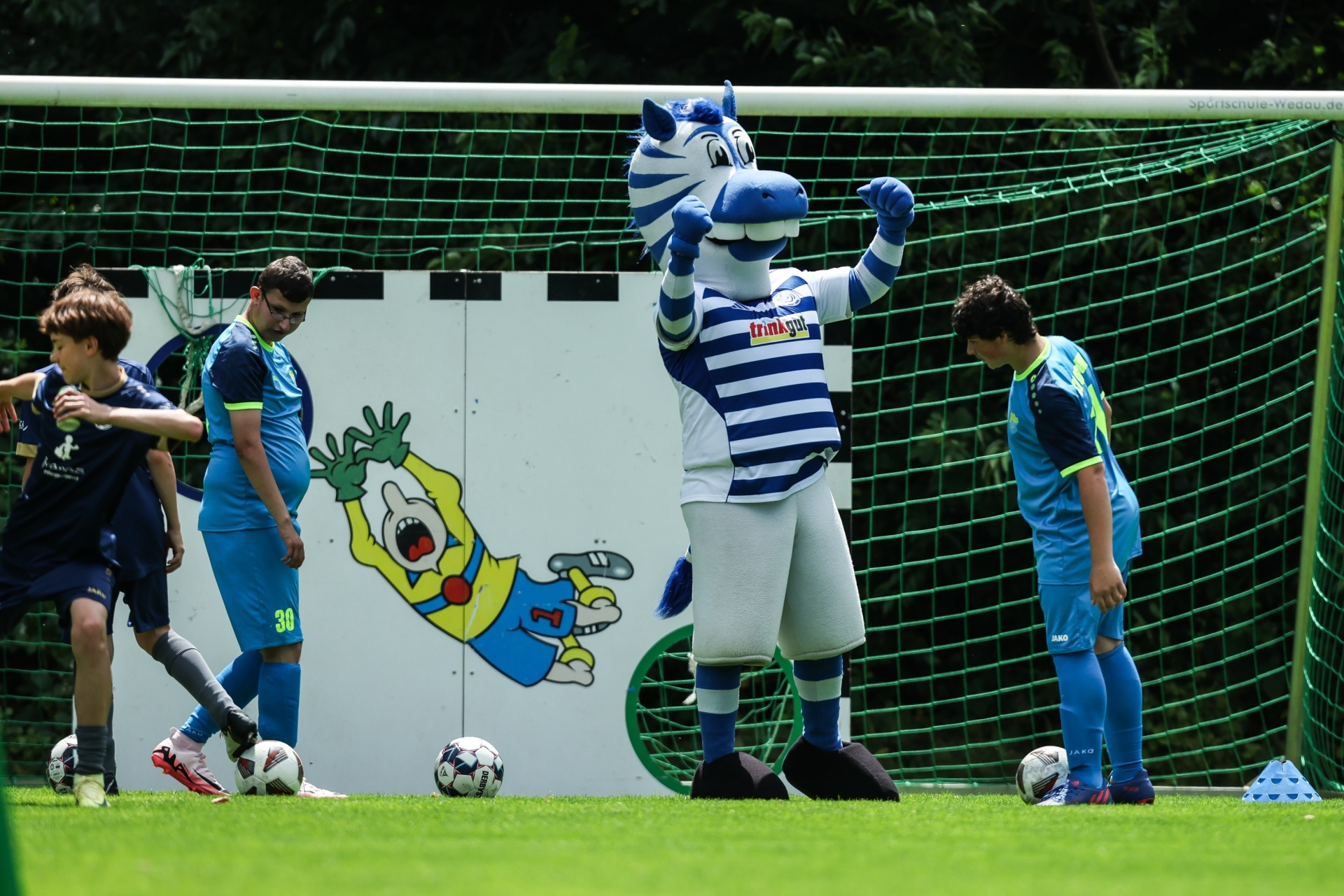 [{"clang_id":1,"value":"Fußballtraining: Vier Jungen mit Fußbällen und ein schwarz-weiß gestreiftes Vereinsmaskottchen (Zebra) auf einem Fußballfeld vor einem Fußballtor mit Wandbemalung."}]