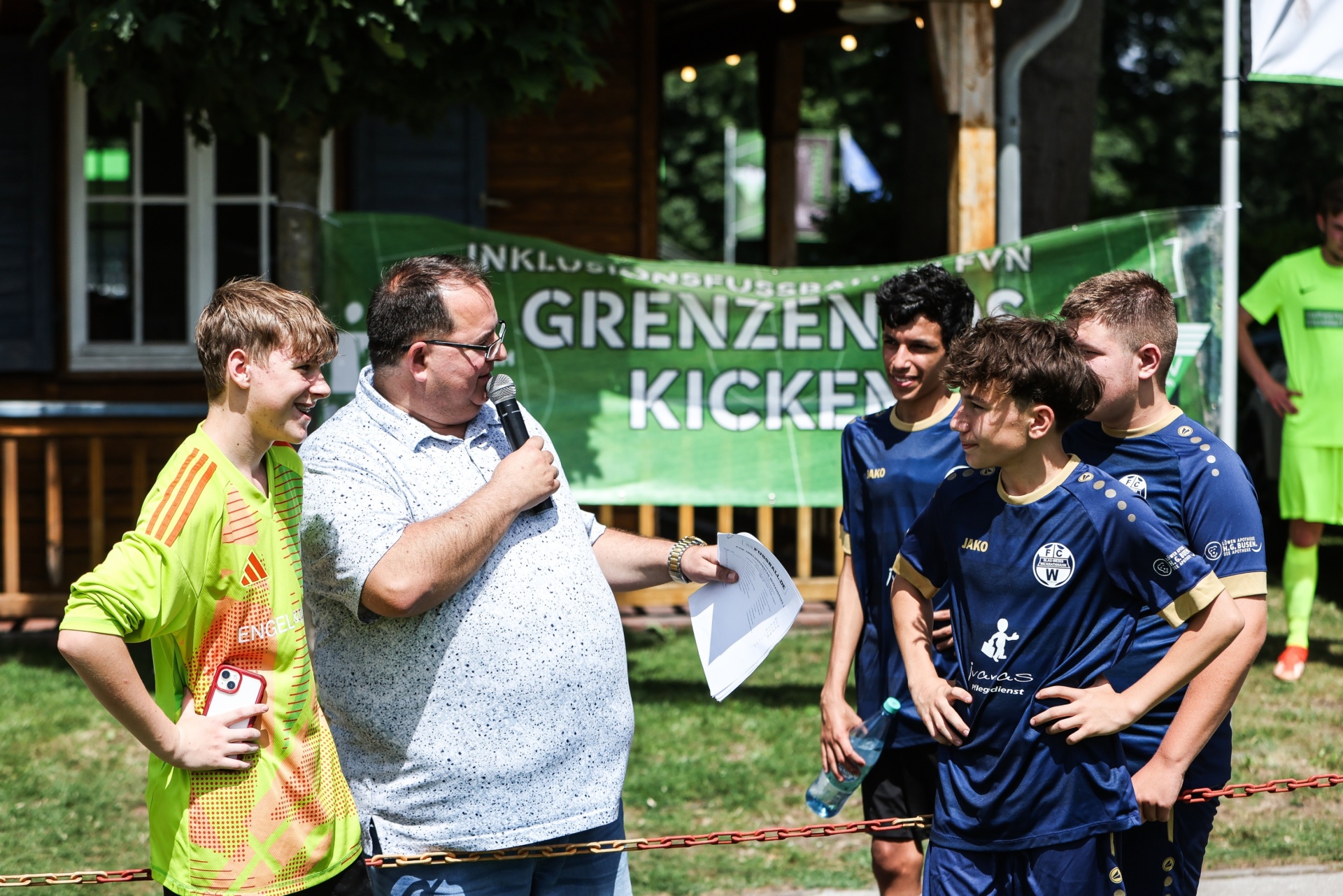 [{"clang_id":1,"value":"Mann mit Mikrofon interviewt Jugendlichen. Im Hintergrund zwei weitere Jugendliche in Fußballtrikots und ein Banner mit Schrift. Outdoor-Szene."}]