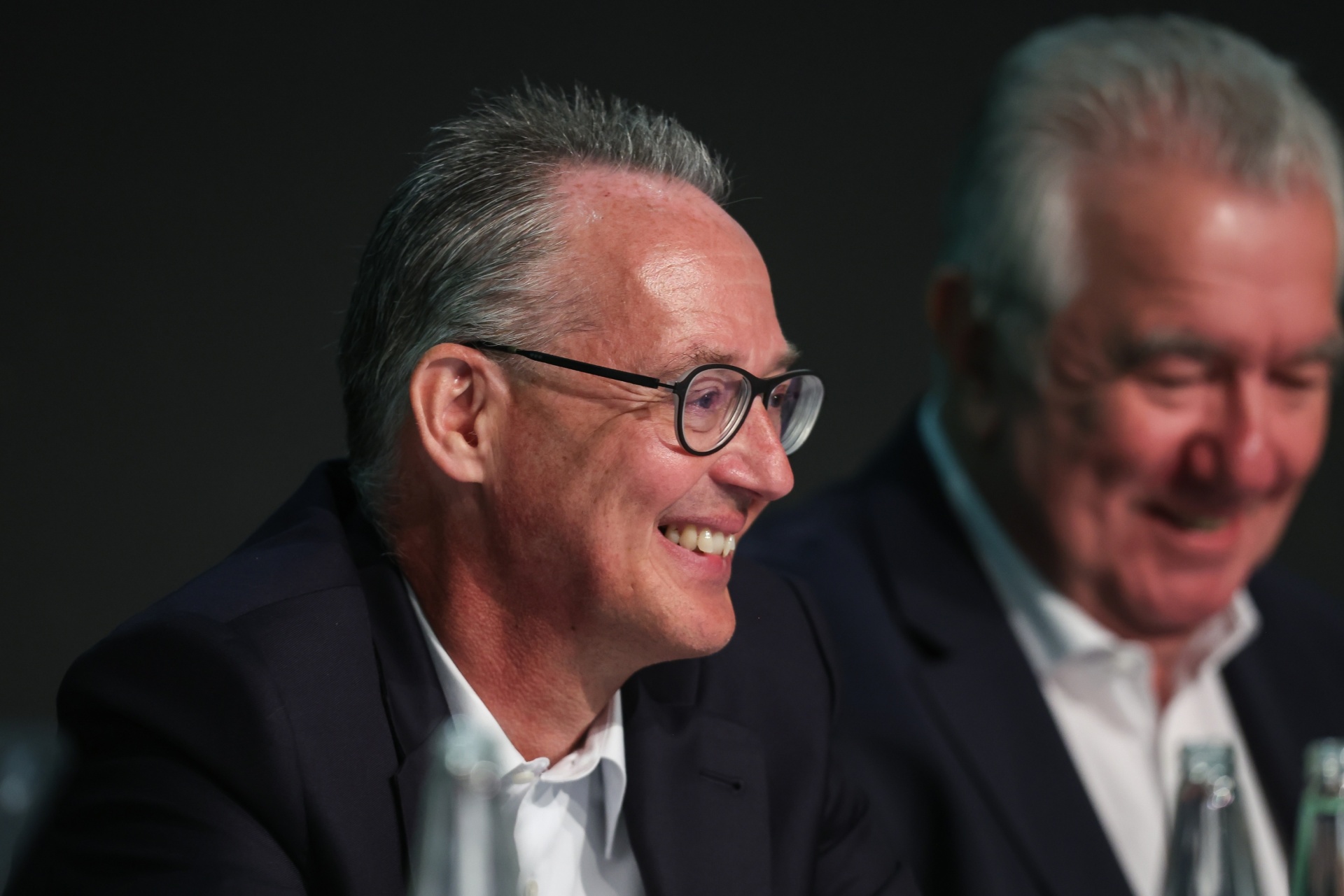 [{"clang_id":1,"value":"Zwei Männer in Anzügen sitzen nebeneinander. Der Mann links trägt eine Brille und lächelt. Der Hintergrund ist dunkel."}]