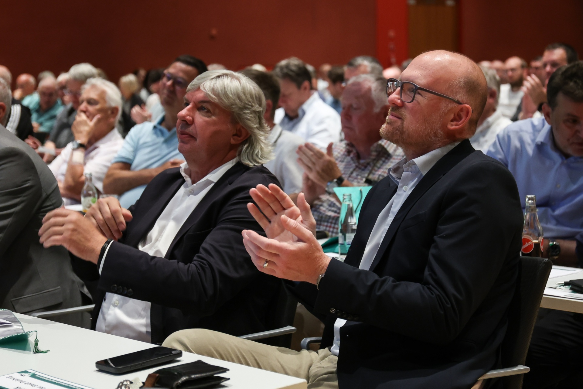 [{"clang_id":1,"value":"Publikum bei einer Veranstaltung. Zwei Männer im Vordergrund klatschen, dahinter weitere Personen in einem Konferenzraum."}]