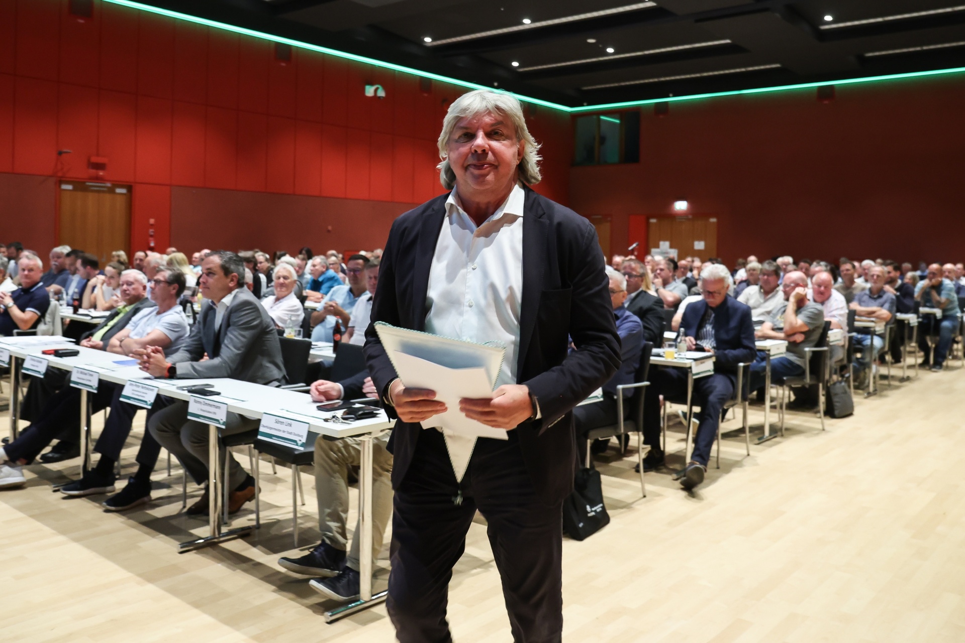 [{"clang_id":1,"value":"Mann mit Jacket und Hemd steht vor einer großen Versammlung von Menschen, die an Tischen in einem Konferenzraum sitzen."}]