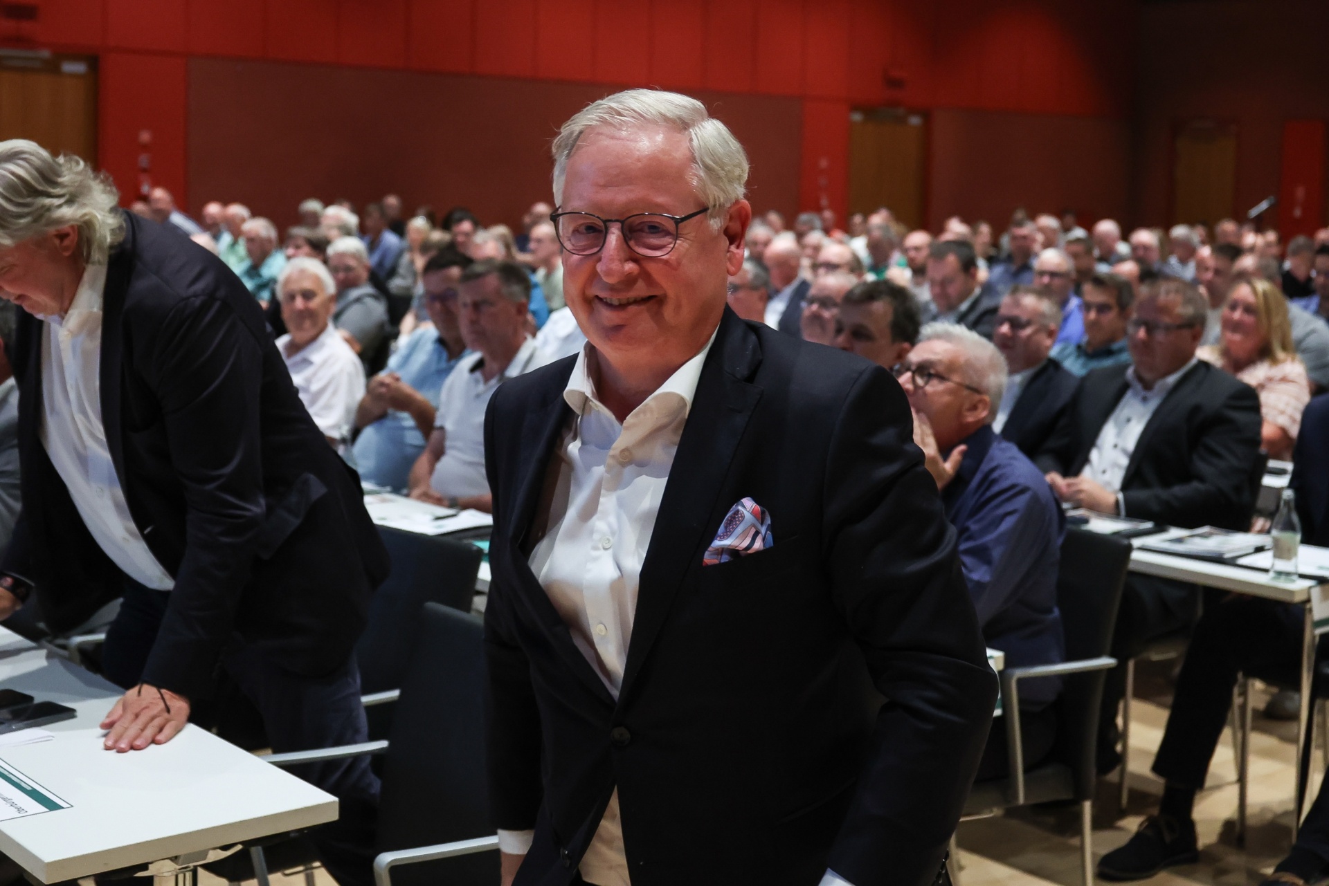 [{"clang_id":1,"value":"Mann im Anzug mit Brille lächelt in einem Konferenzraum. Im Hintergrund viele weitere Personen und Reihen von Tischen."}]