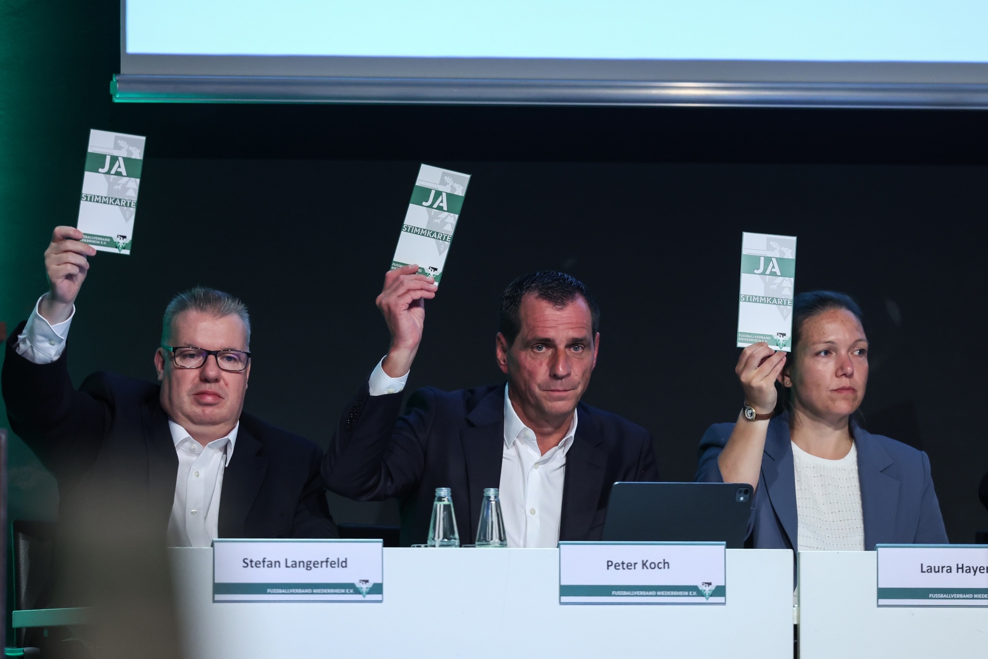 [{"clang_id":1,"value":"Drei Personen sitzen an einem Tisch mit Namensschildern: Stefan Langerfeld, Peter Koch, Laura Hayen. Alle drei halten grüne Karten mit dem Aufdruck \"JA\" hoch."}]