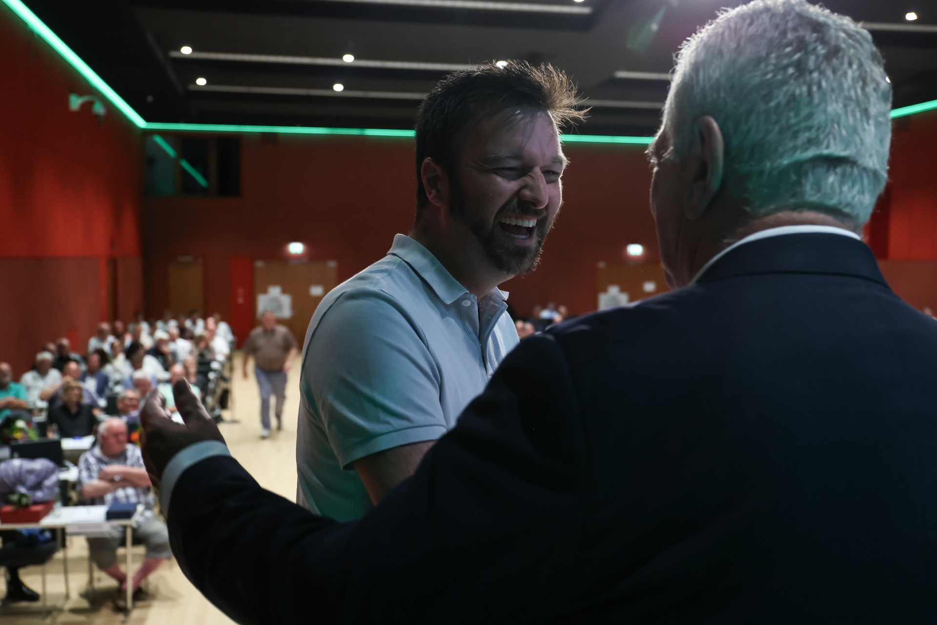 [{"clang_id":1,"value":"Zwei Männer lächeln und unterhalten sich in einer großen Halle mit vielen Personen im Hintergrund. Der eine trägt Anzug, der andere ein hellblaues Hemd."}]
