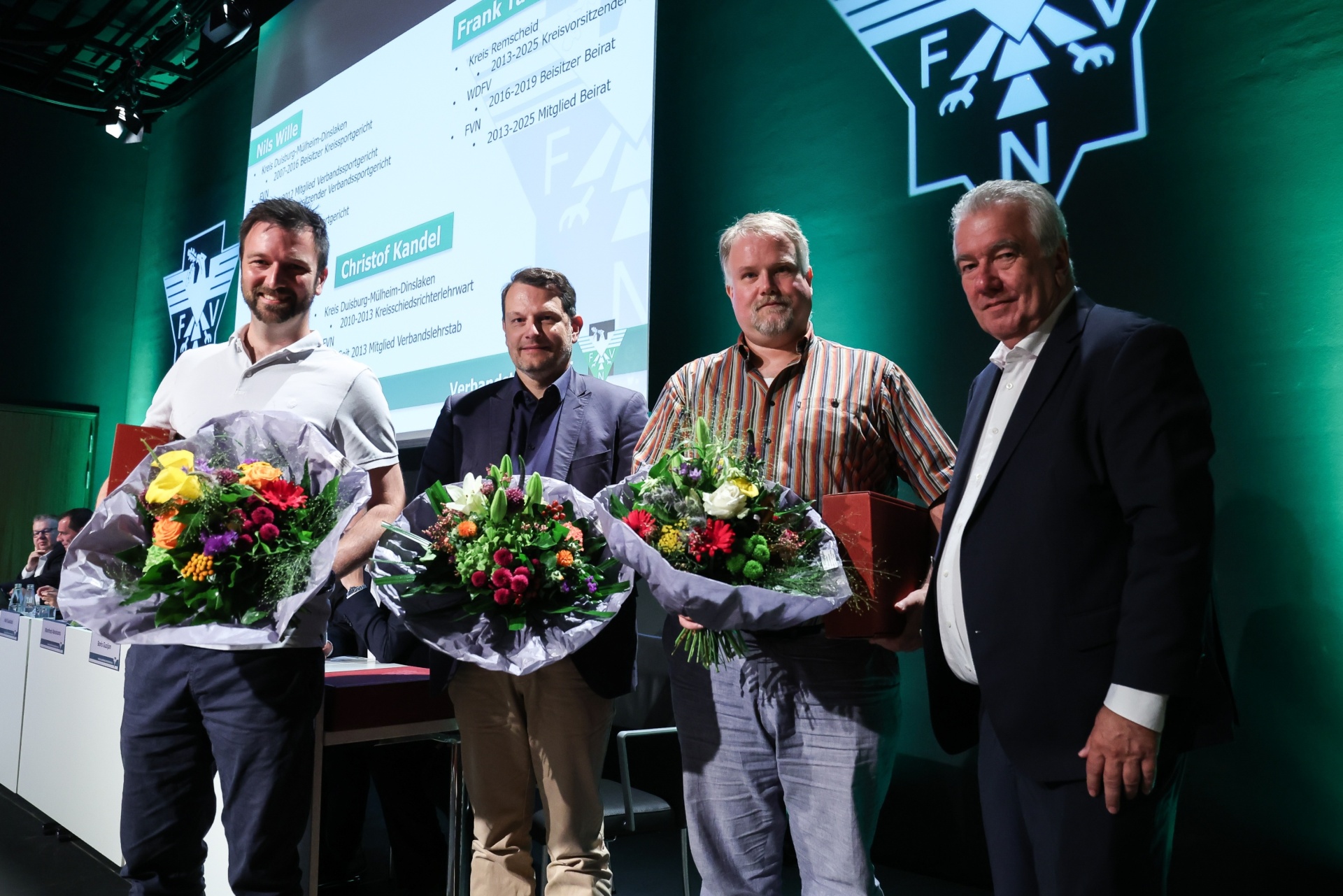 [{"clang_id":1,"value":"Fünf Männer stehen auf einer Bühne und halten Blumensträuße. Im Hintergrund eine grüne Wand mit Logos und Text."}]