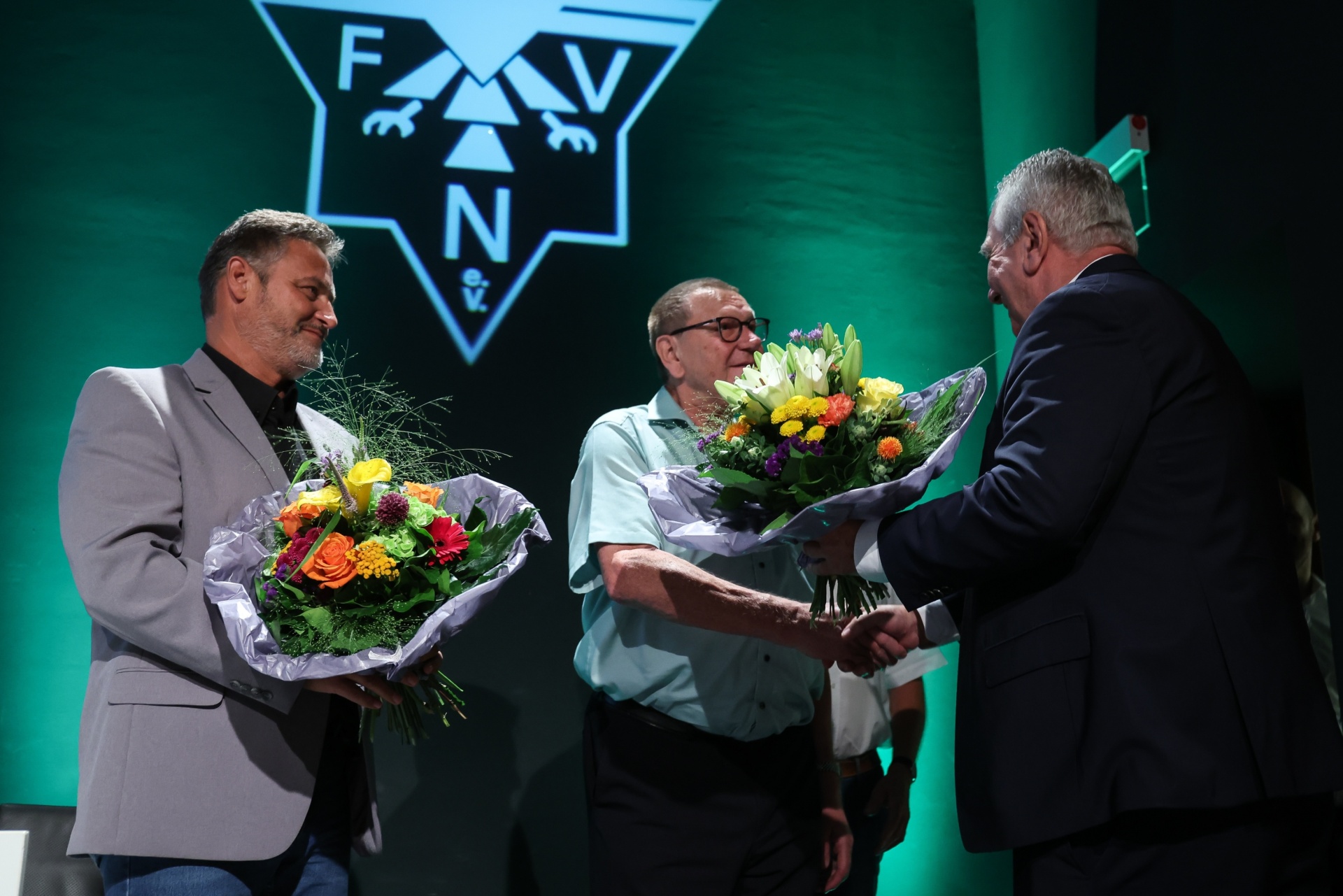 [{"clang_id":1,"value":"Drei Männer auf einer Bühne. Zwei erhalten Blumensträuße vom dritten Mann. Im Hintergrund ein großes grünes Logo mit den Buchstaben F, N, V."}]