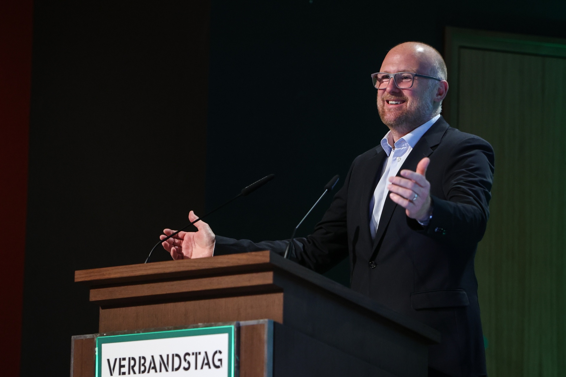 [{"clang_id":1,"value":"Mann mit Glatze in schwarzem Anzug spricht an einem Pult. Auf dem Pult steht \"VERBANDSTAG\" in weißer Schrift."}]