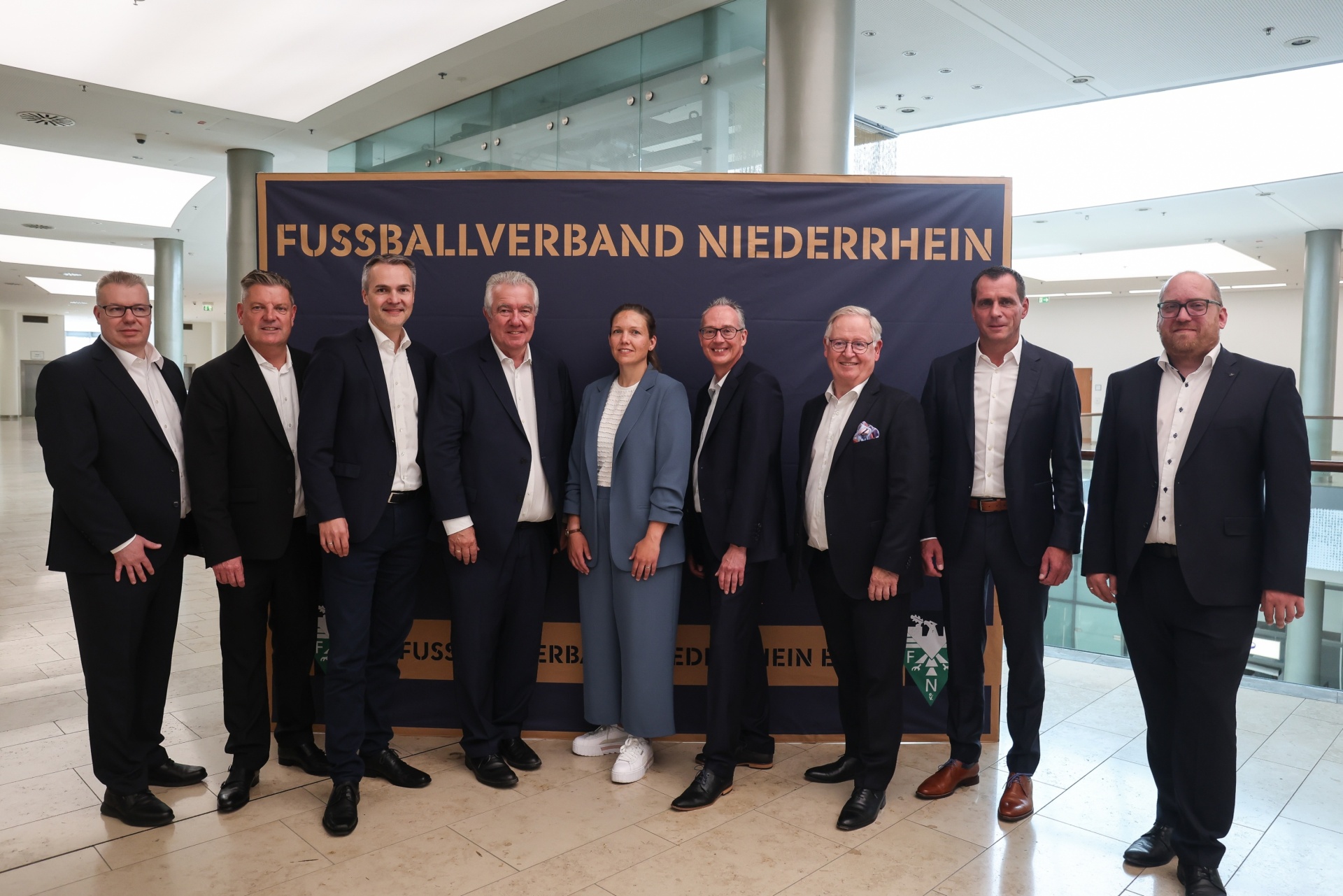 [{"clang_id":1,"value":"Gruppe von neun Personen in Anzügen vor Banner mit Aufschrift \"FUSSBALLVERBAND NIEDERRHEIN\". Helle Lobby mit Marmorboden."}]