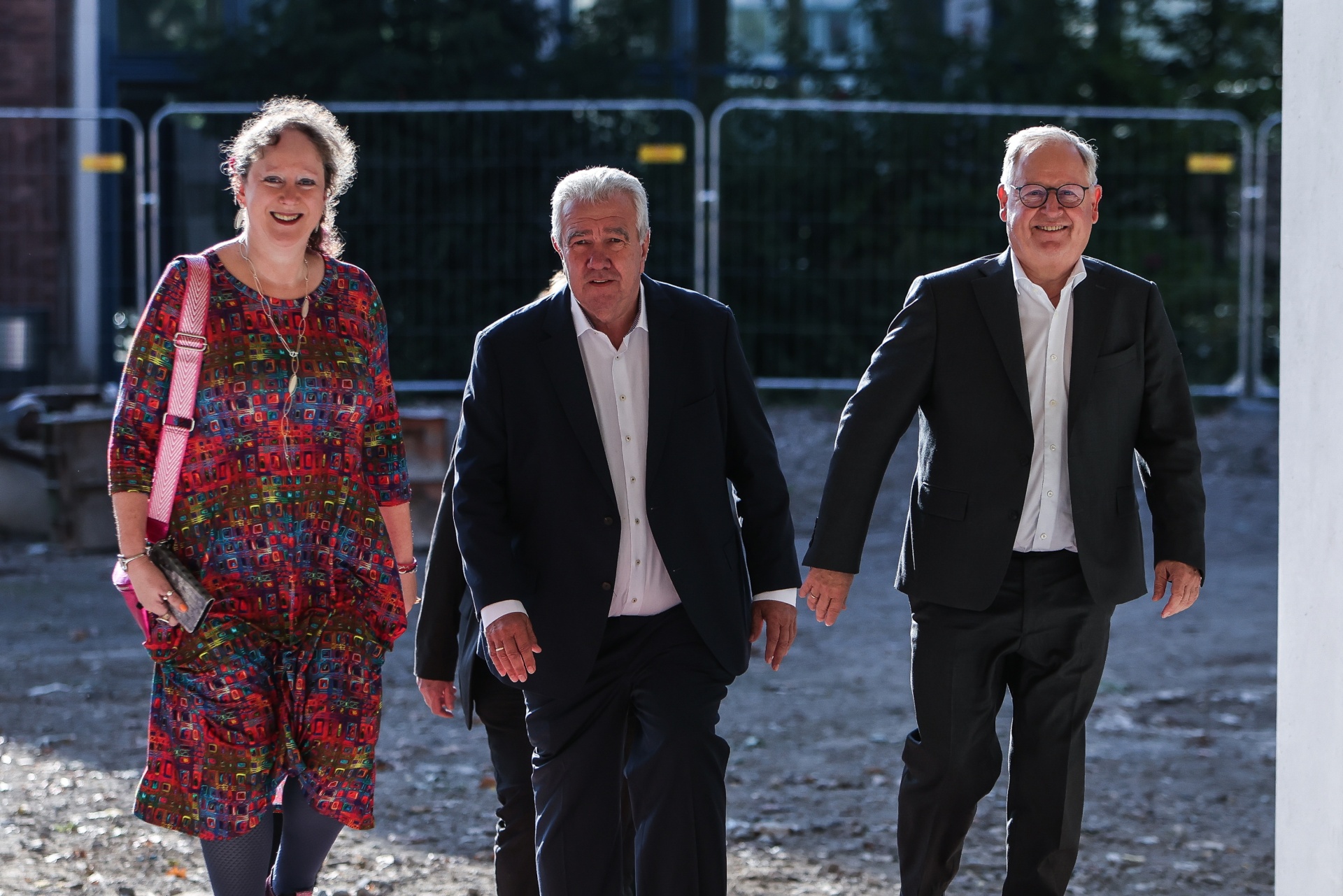 [{"clang_id":1,"value":"Drei Personen gehen nebeneinander. Links eine Frau mit gemustertem Kleid, in der Mitte und rechts Männer in dunklen Anzügen. Im Hintergrund Zäune und Gebäude."}]