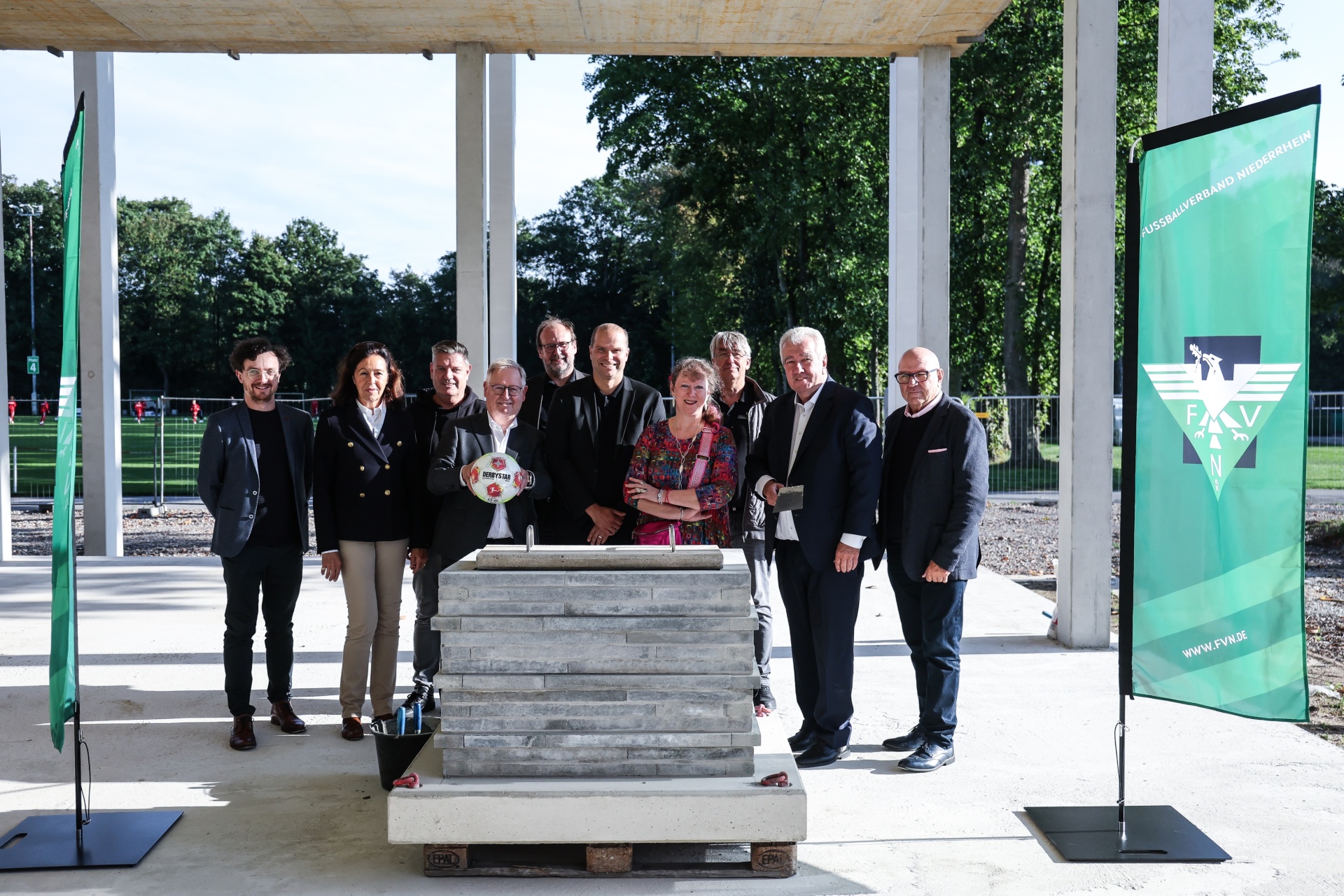 [{"clang_id":1,"value":"Gruppe von neun Personen steht um eine graue Steininstallation auf einer Betonfläche unter einem weißen Pavillon. Im Hintergrund ein Fußballfeld und Bäume."}]