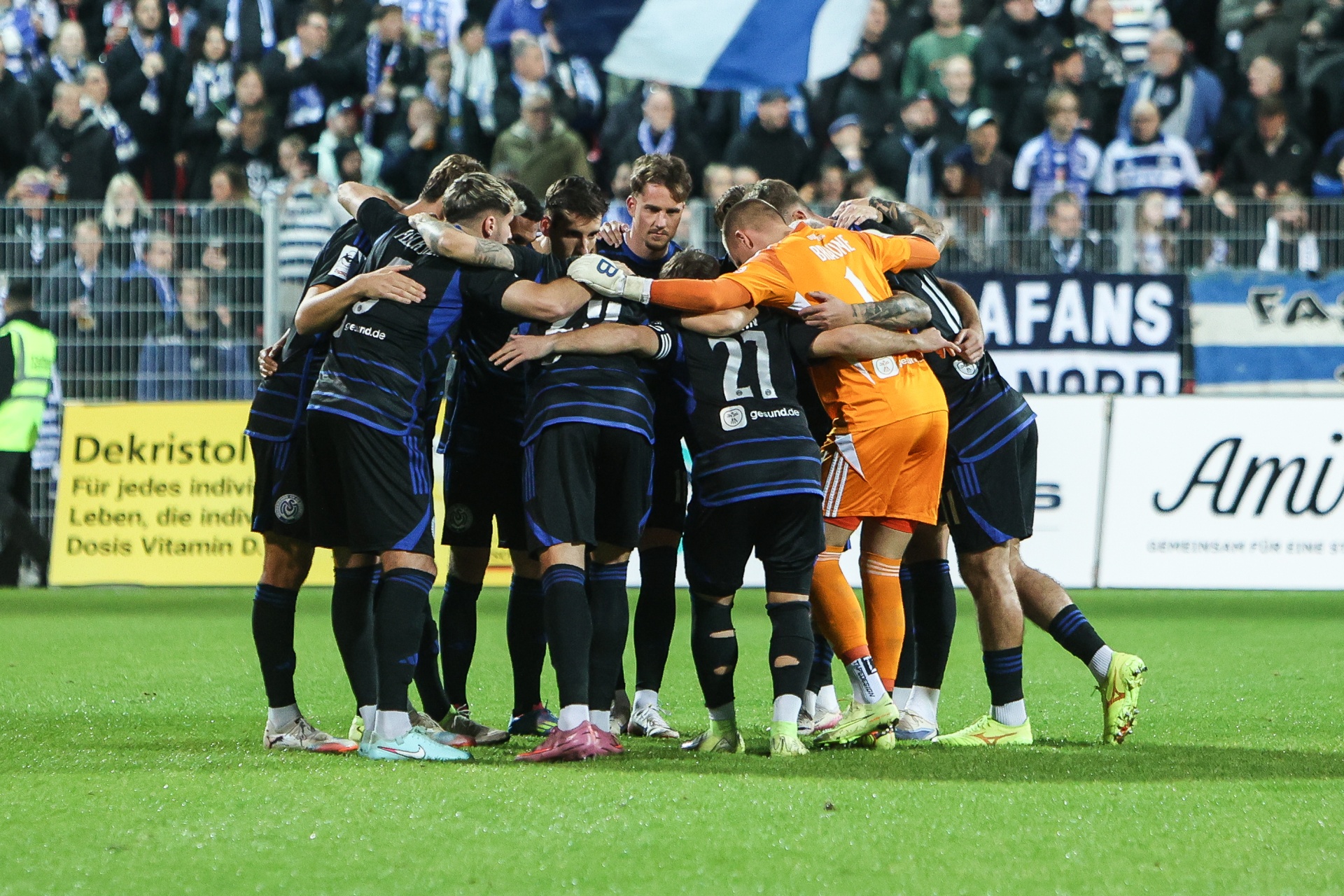 [{"clang_id":1,"value":"Fußballspieler in schwarz-blauen Trikots und orangefarbenen Torwarttrikots feiern eng umschlungen auf grünem Rasen. Im Hintergrund Fans auf den Tribünen."}]