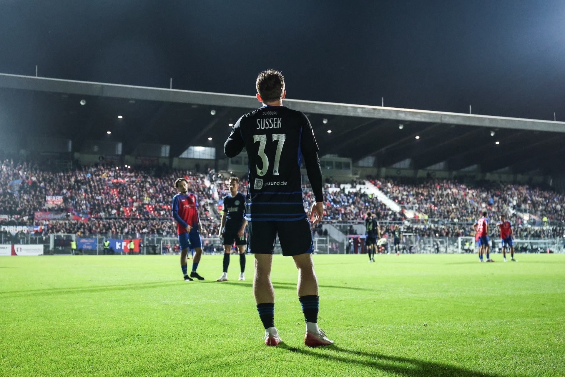 [{"clang_id":1,"value":"Fußballspieler mit Rückennummer 37 auf grünem Rasen, Arm hoch, vor gefülltem Stadion bei Nacht. Gegenüber stehen gegnerische Spieler."}]