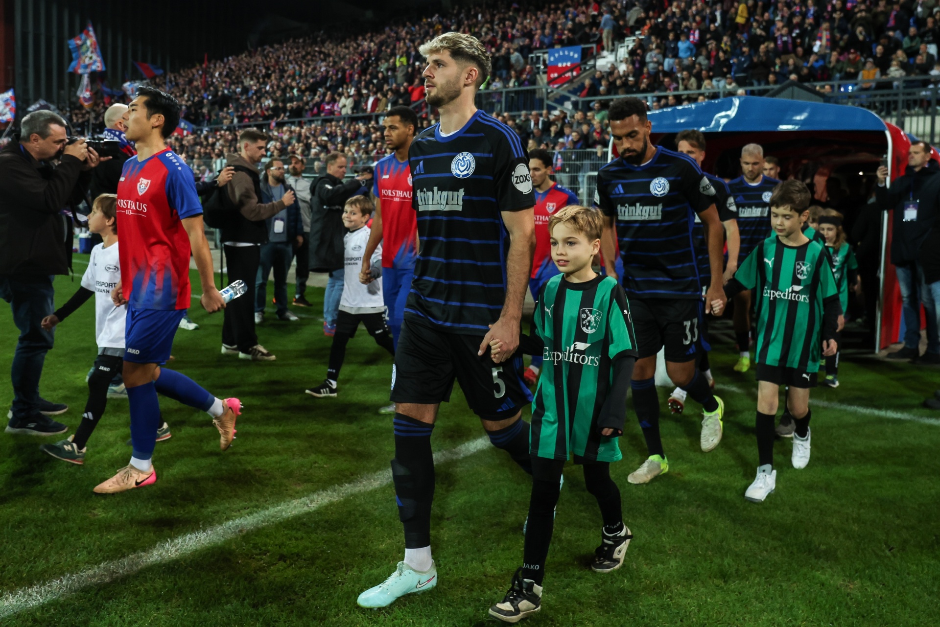 [{"clang_id":1,"value":"Fußballspieler und Kinder in Vereinskleidung betreten einen Fußballplatz. Im Hintergrund sind Zuschauer auf den Tribünen zu sehen."}]