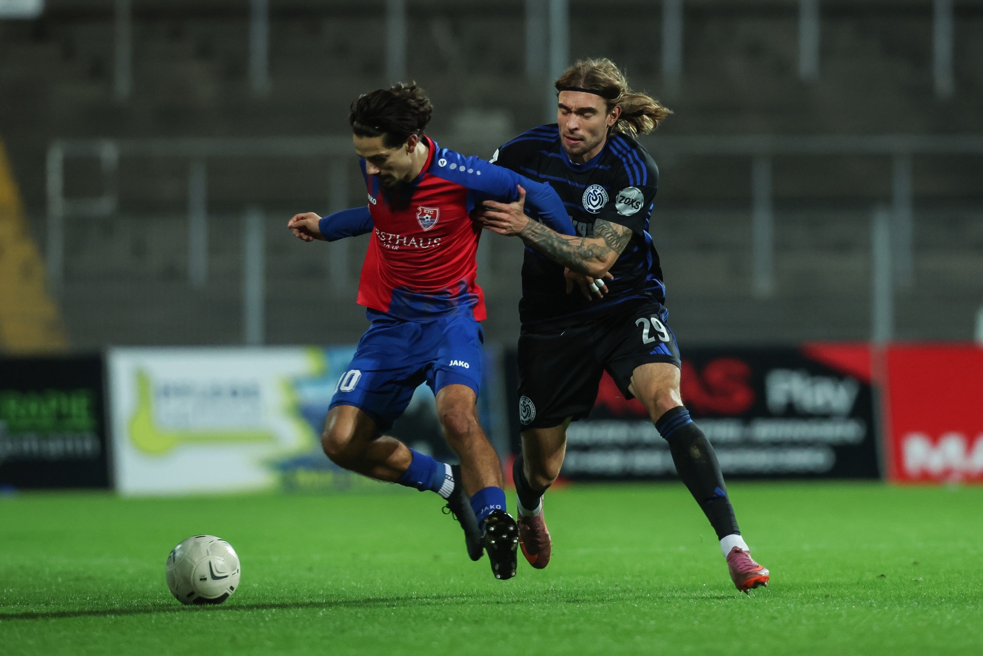 [{"clang_id":1,"value":"Zwei Fußballspieler im Zweikampf auf grünem Rasen. Links rotes Trikot mit Nummer 10, rechts schwarzes Trikot mit Nummer 29. Im Hintergrund ein Stadion."}]