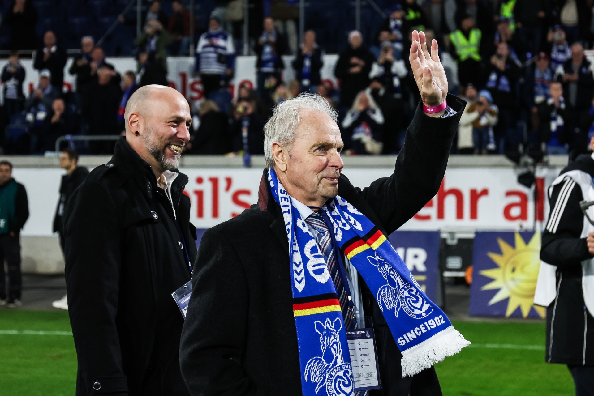 [{"clang_id":1,"value":"Zwei Männer auf einem Fußballfeld. Der ältere Mann im Vordergrund trägt einen blauen Schal und winkt. Im Hintergrund sind Zuschauerreihen zu sehen."}]