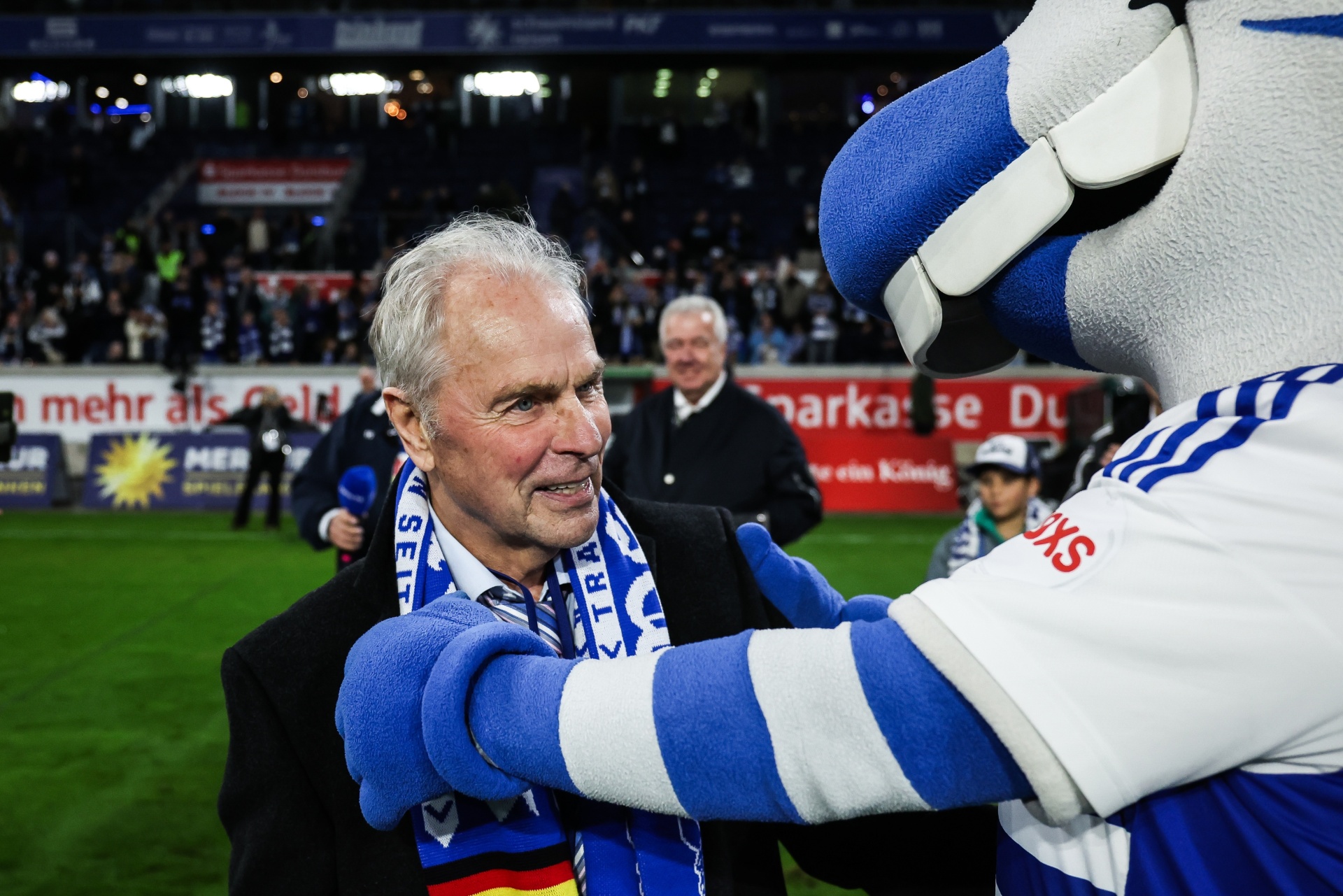 [{"clang_id":1,"value":"Mann mit blau-weißem Schal wird von einem blauen Maskottchen umarmt. Im Hintergrund Fußballstadion mit Zuschauern."}]