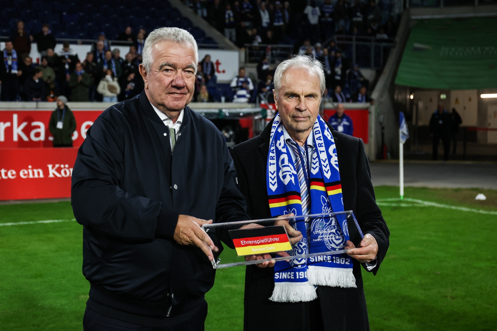 [{"clang_id":1,"value":"Zwei ältere Männer stehen auf einem Fußballfeld. Der linke trägt eine dunkle Jacke, der rechte ein blau-weißes Vereinsoutfit und hält eine Auszeichnung entgegen. Im Hintergrund Zuschauer."}]