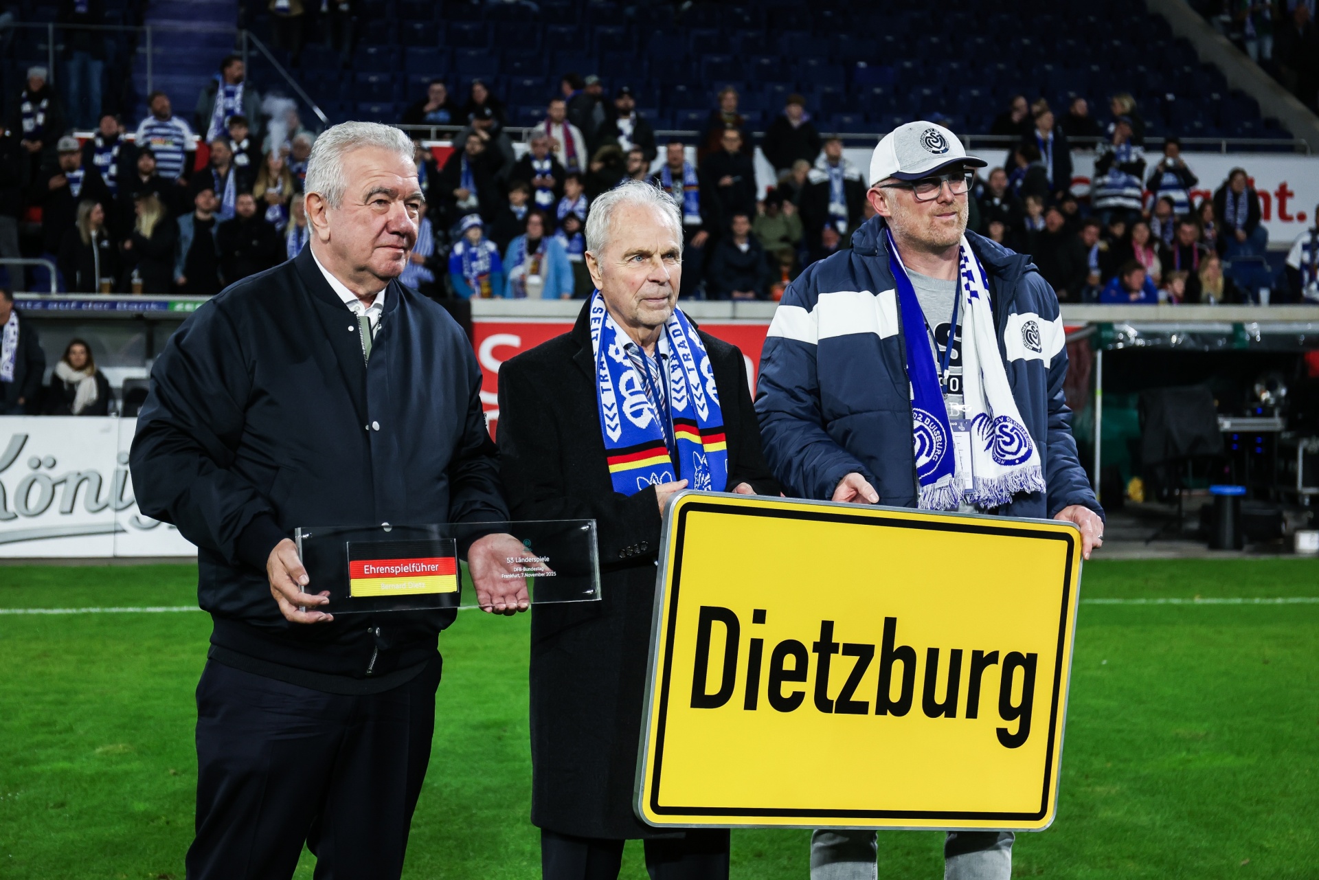 [{"clang_id":1,"value":"Drei Männer stehen auf einem Fußballfeld vor Zuschauern. Sie halten schwarze Plaketten und ein Straßenschild mit der Aufschrift \"Dietzburg\"."}]