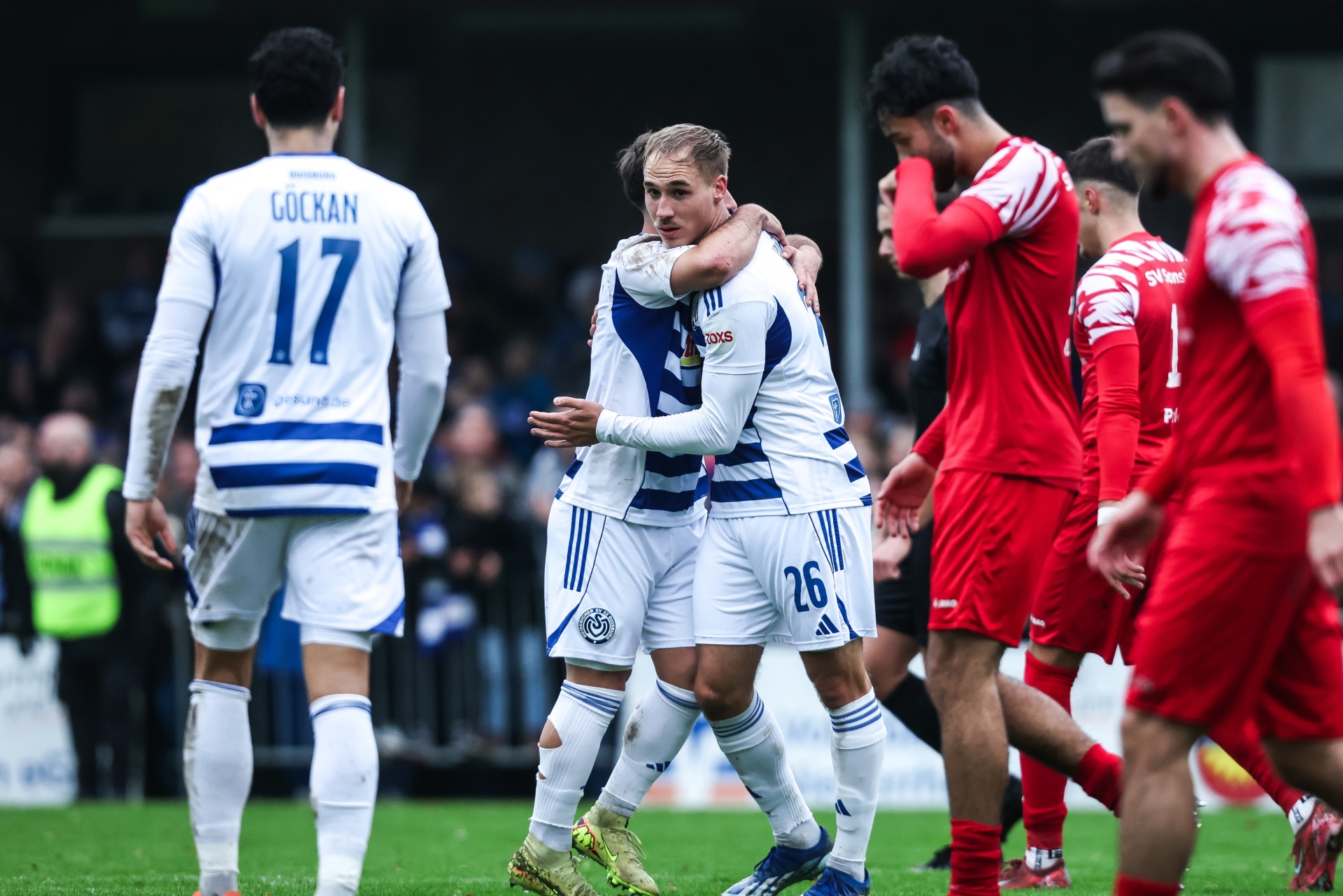 [{"clang_id":1,"value":"Zwei Fußballspieler in blau-weißen Trikots umarmen sich auf dem Spielfeld. Im Hintergrund stehen weitere Spieler in roten und weißen Trikots. Rasen und Zuschauer im Hintergrund."}]