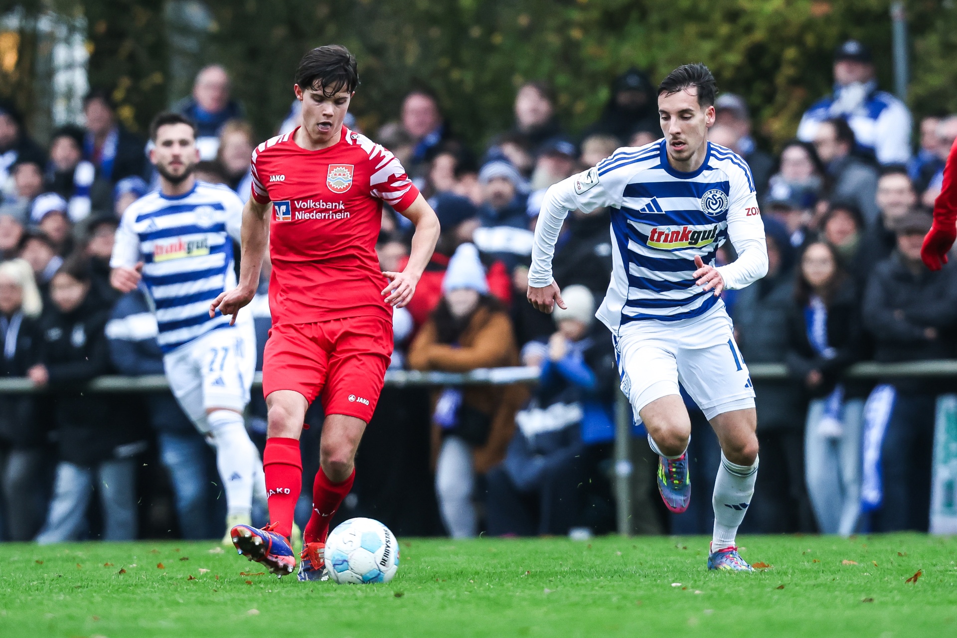 [{"clang_id":1,"value":"Zwei Fußballspieler im Wettkampf auf Grün. Links rotes Trikot, rechts blau-weiß gestreiftes. Zuschauer im Hintergrund."}]