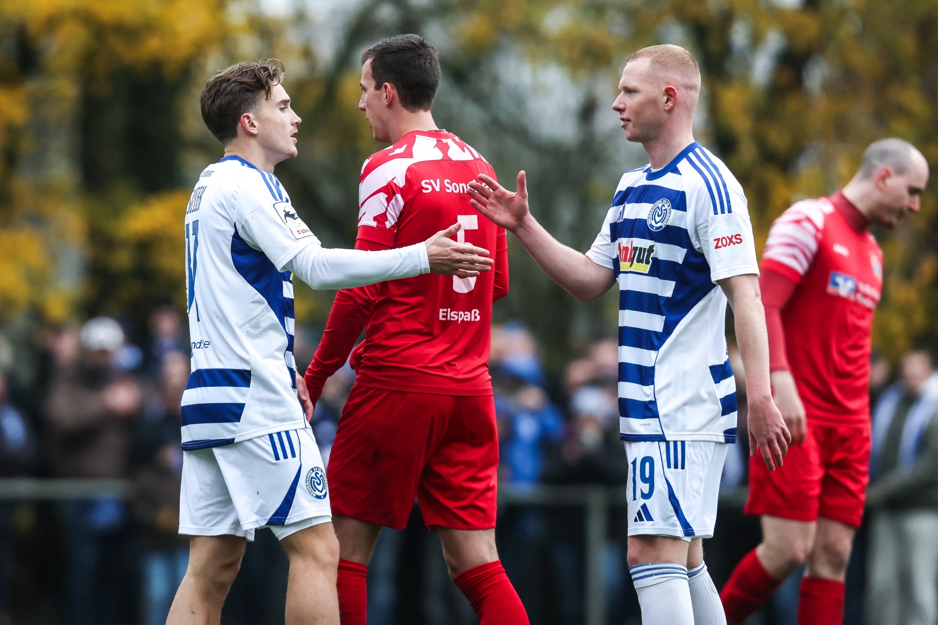 [{"clang_id":1,"value":"Zwei Fußballspieler im rot-weiß gestreiften Trikot und ein Spieler in Rot stehen sich auf dem Feld gegenüber. Im Hintergrund Zuschauer und Bäume."}]