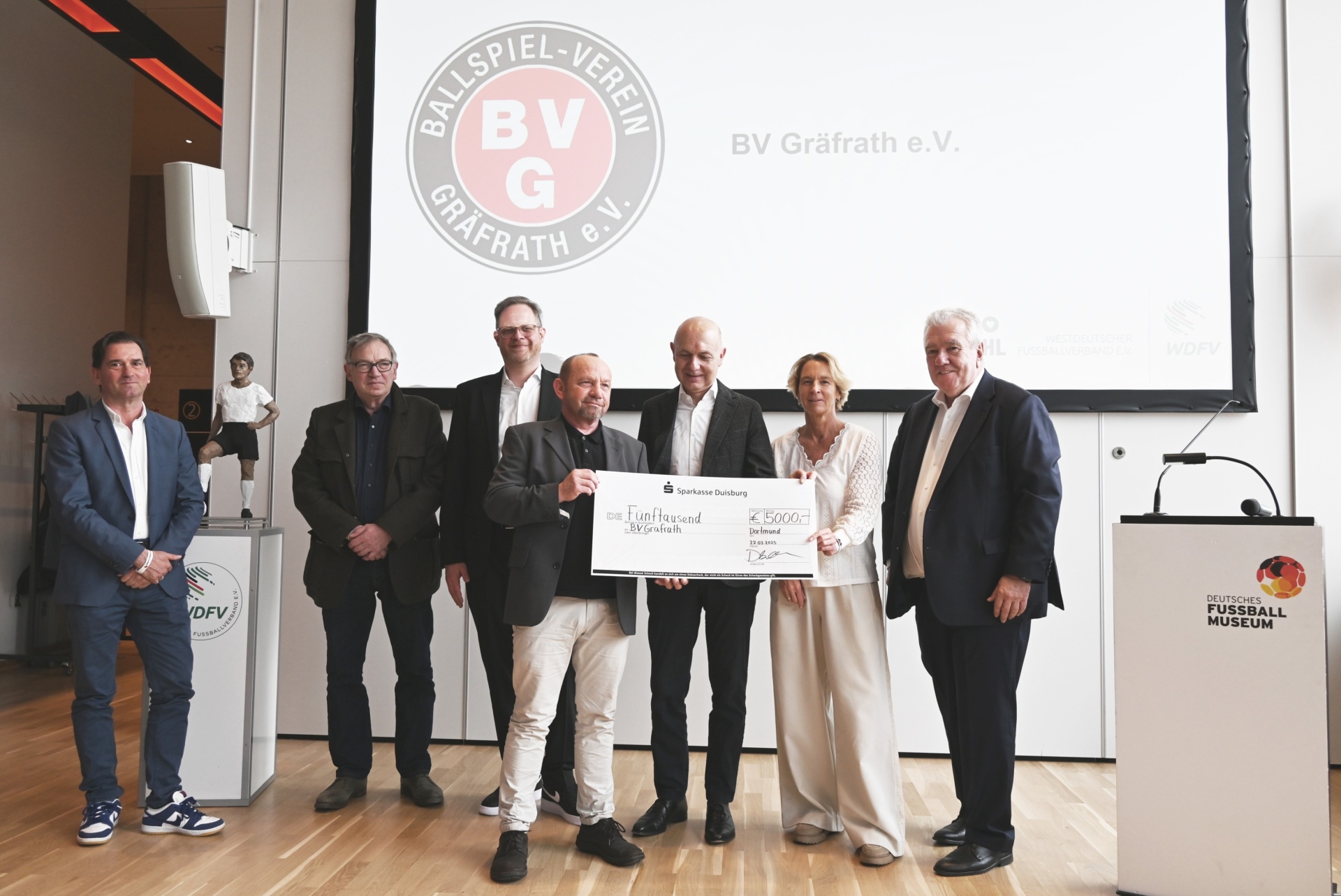[{"clang_id":1,"value":"Gruppe posiert vor Logo \"BV Gräfrath e.V.\". Ein Mann übergibt einen symbolischen Scheck. Im Hintergrund das Deutsche Fußballmuseum."}]