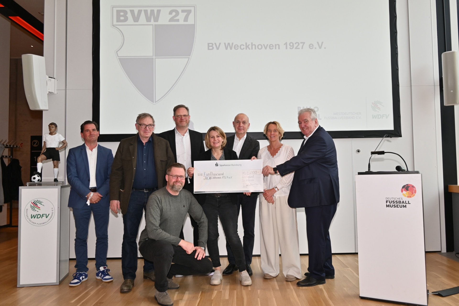 [{"clang_id":1,"value":"Gruppe von neun Personen bei einer Preisübergabe. Im Hintergrund ein Banner mit dem Schriftzug \"BVW 27\" und \"BV Weckhoven 1927 e.V.\". Zwei Rednerpulte mit Logo \"WDFV\" und \"Deutsches Fußballmuseum\"."}]