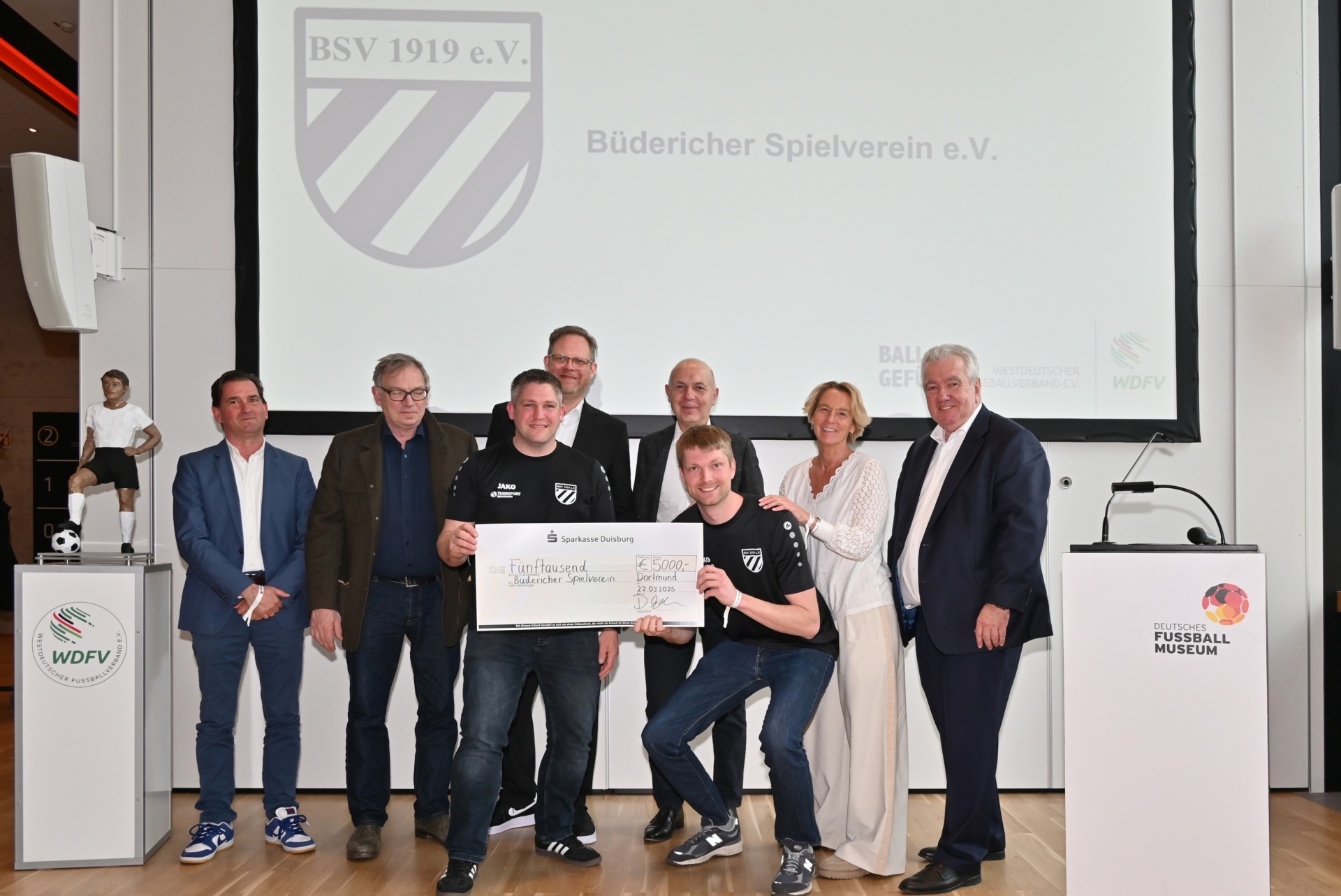 [{"clang_id":1,"value":"Gruppe vor Banner \"BSV 1919 e.V.\". Männer und Frauen halten einen übergroßen Scheck. Im Hintergrund Fußballmuseum-Logo und weitere Banner."}]
