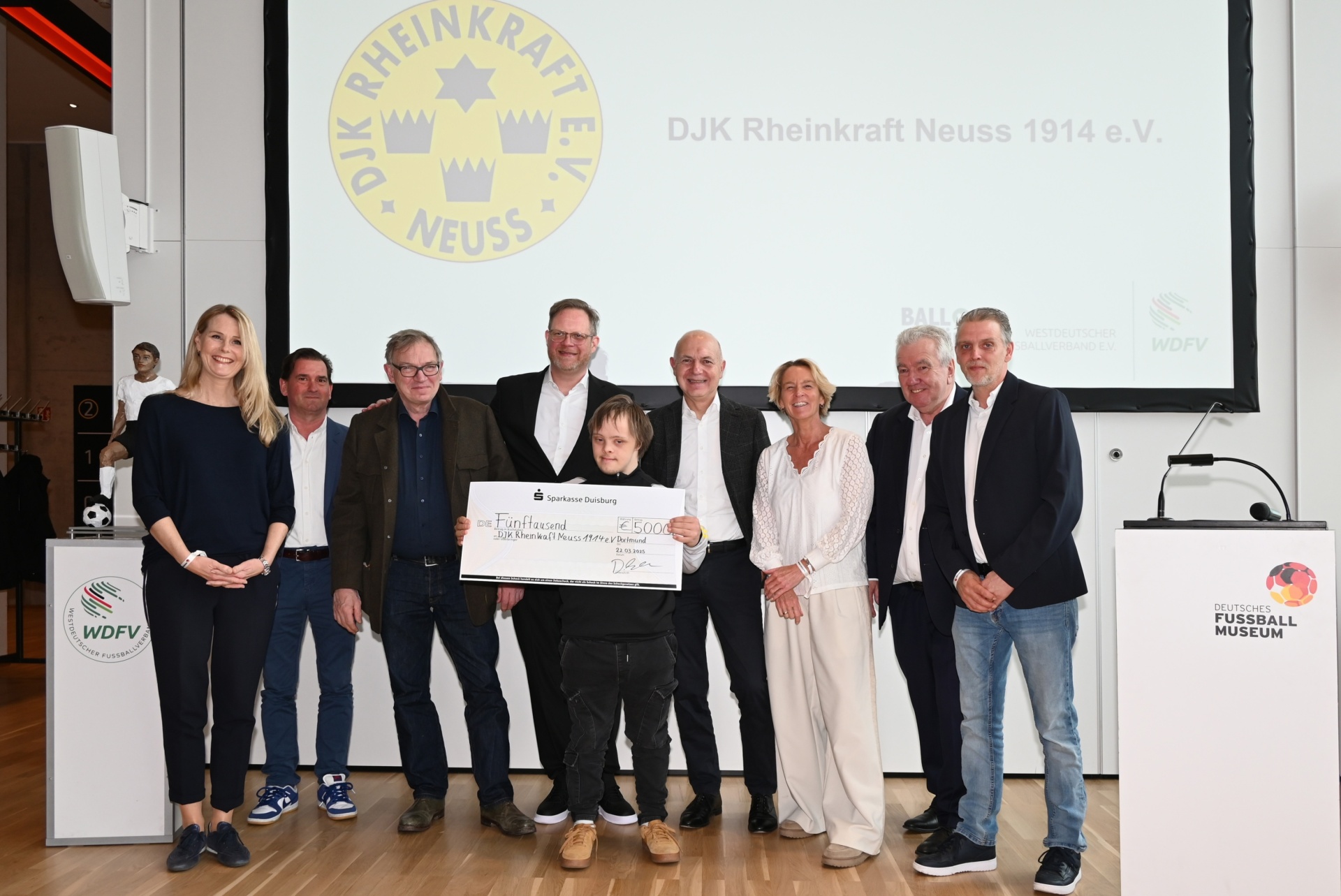 [{"clang_id":1,"value":"Gruppe posiert mit übergroßem Scheck vor Logo \"DJK Rheinkraft Neuss 1914 e.V.\". Im Hintergrund Präsentationstechnik und Logos von Sportverbänden."}]