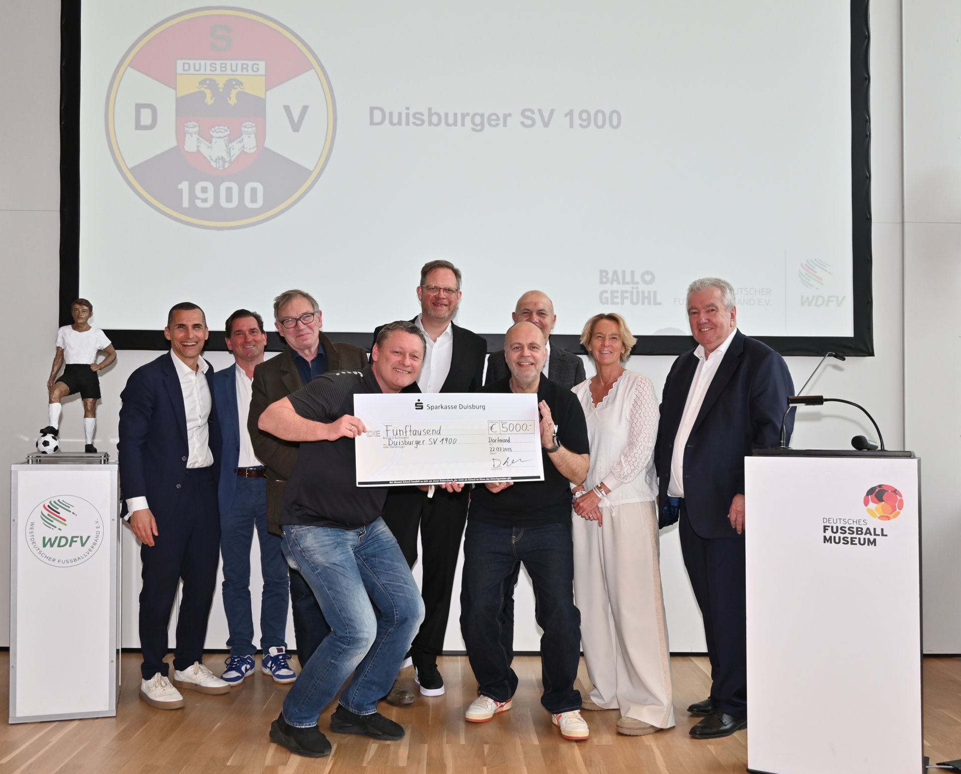 [{"clang_id":1,"value":"Gruppe posiert vor Leinwand mit Vereinslogo \"Duisburger SV 1900\". Ein Mann übergibt einen symbolischen Scheck, weitere Personen stehen darum herum."}]