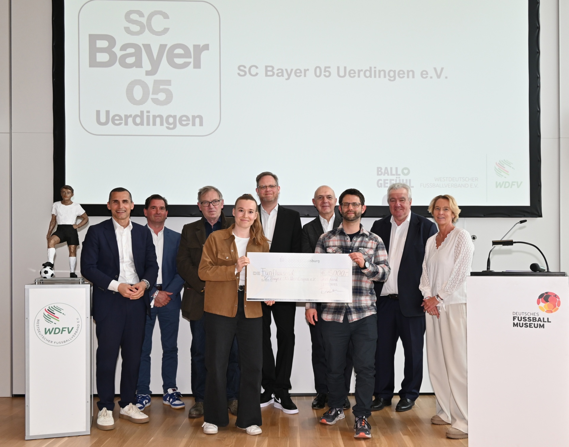 [{"clang_id":1,"value":"Gruppe posiert mit übergroßem Scheck vor Logo \"SC Bayer 05 Uerdingen\". Hintergrund: Ausstellungsraum des Deutschen Fußballmuseums."}]
