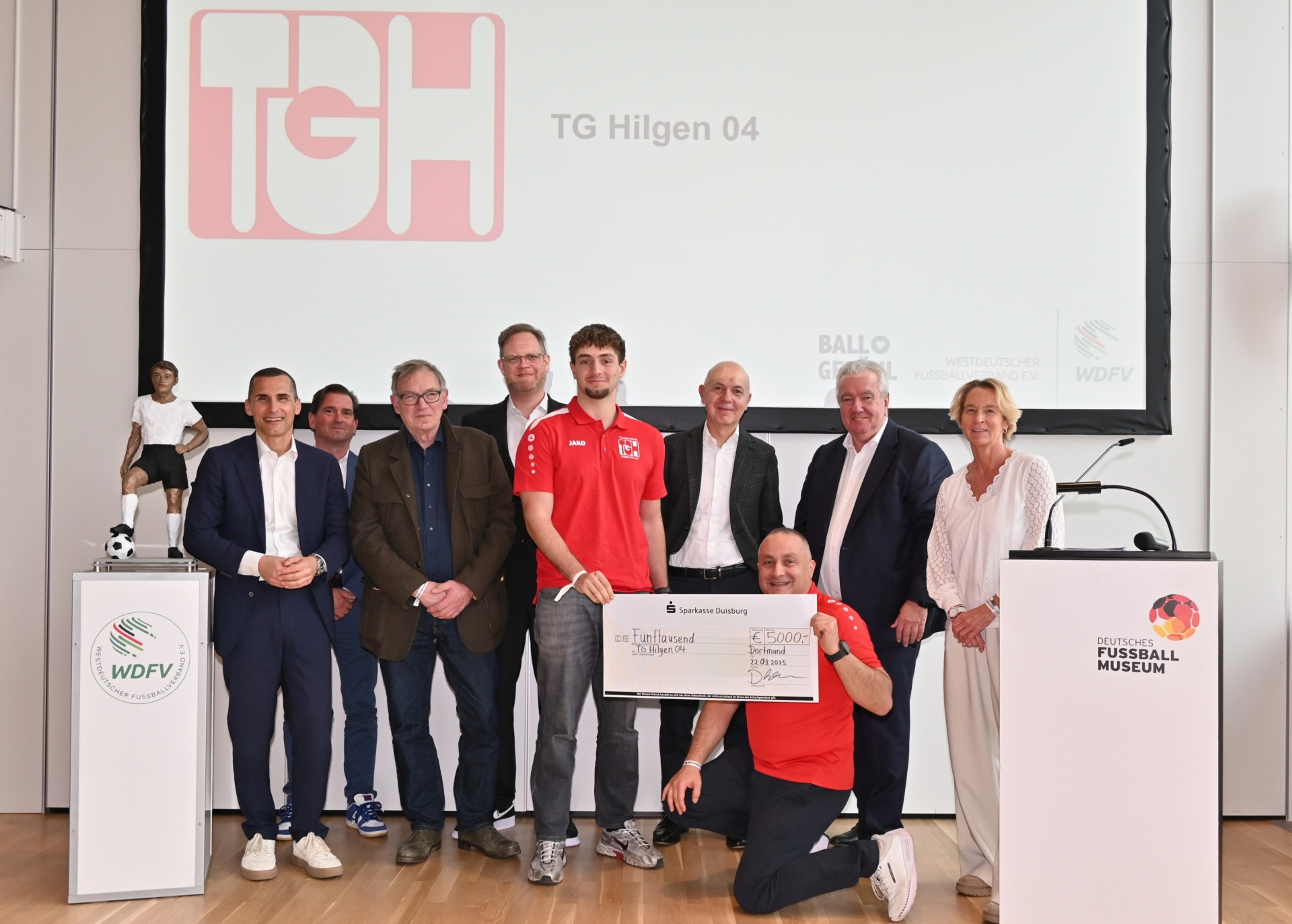 [{"clang_id":1,"value":"Gruppe posiert mit übergroßen Scheck. Im Hintergrund Logo \"TG Hilgen 04\" und Wappen. Eine Person kniet mit rotem T-Shirt davor."}]
